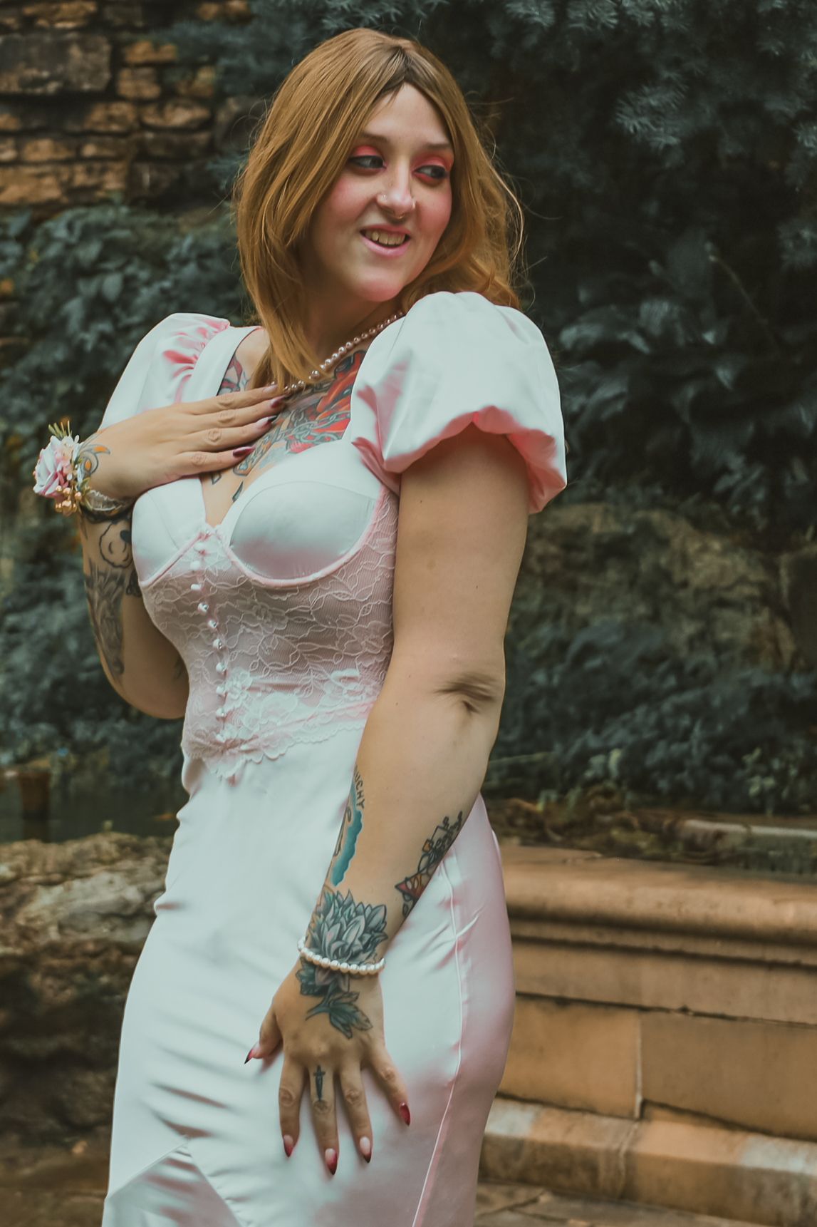 A person in a white lace dress poses outdoors against a stone wall showing off their tattoo design.