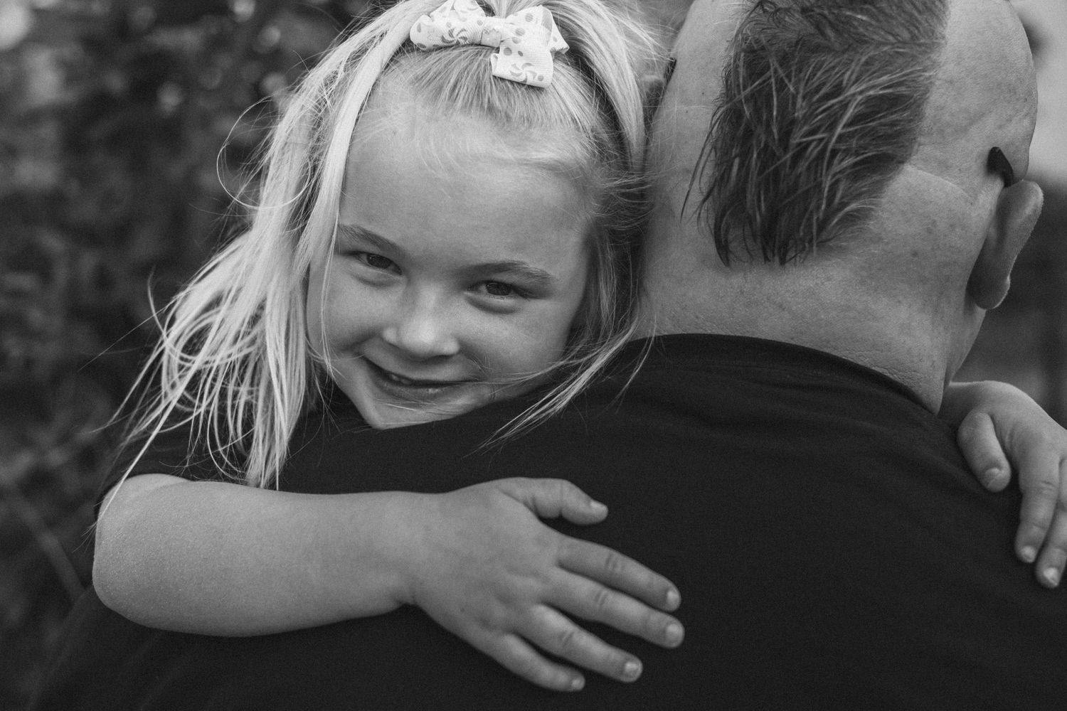 A young blonde girl with a bow hugs a man from behind, smiling warmly in a black and white portrait photo.