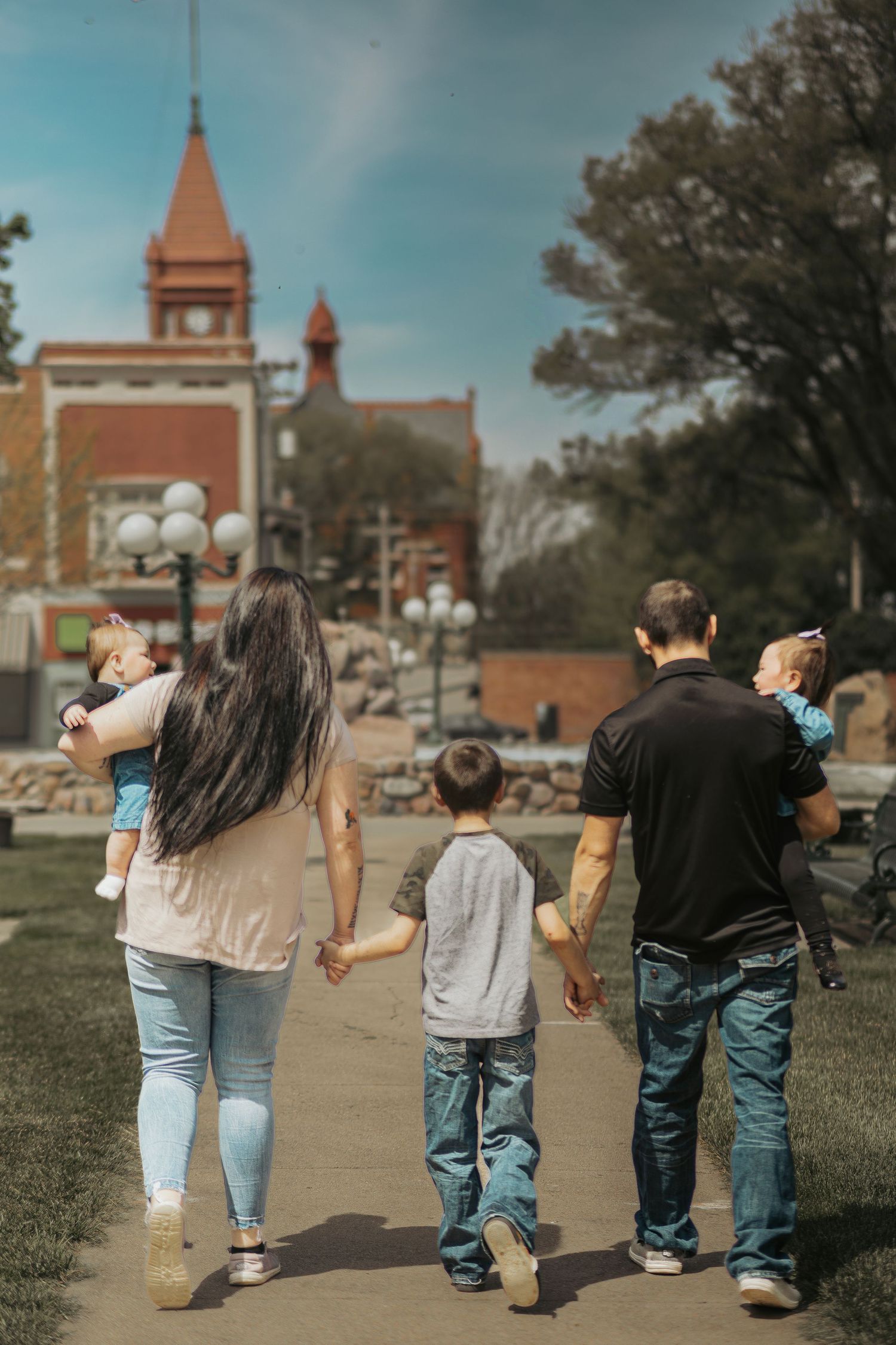 Family of five walking hand-in-hand through a park toward a historic red brick building on a sunny day.