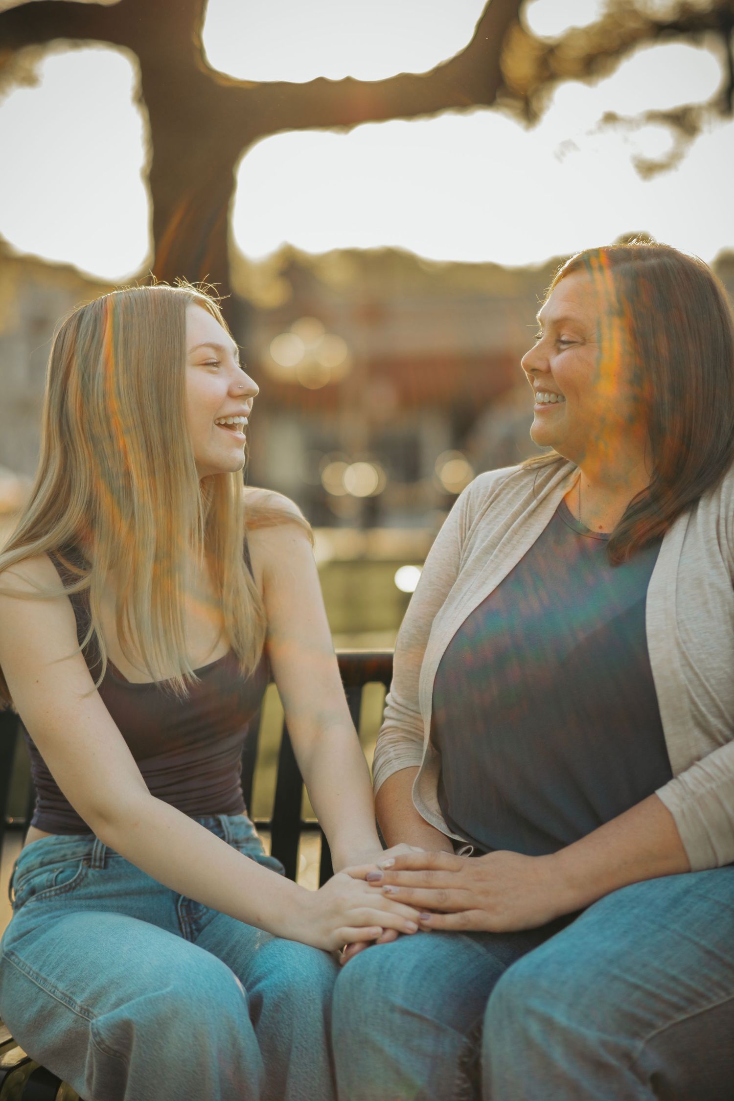 Two women sitting outdoors laughing and holding hands in warm golden sunset light.