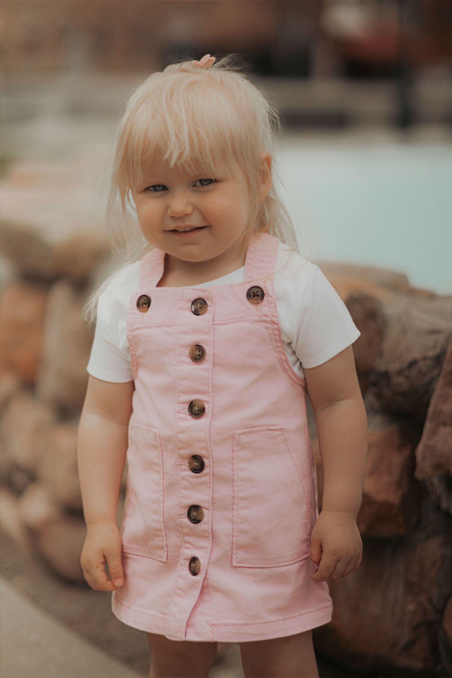 Smiling toddler girl in pink pinafore dress over white tee, with blonde hair and a pink bow.