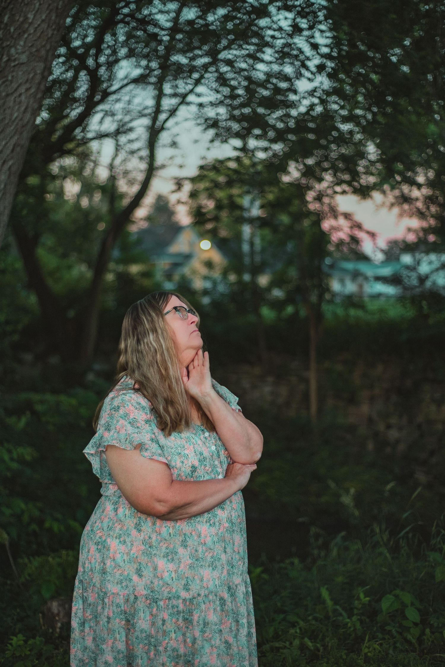 A plus-size woman in a floral dress gazes upward thoughtfully in a lush green garden at dusk.