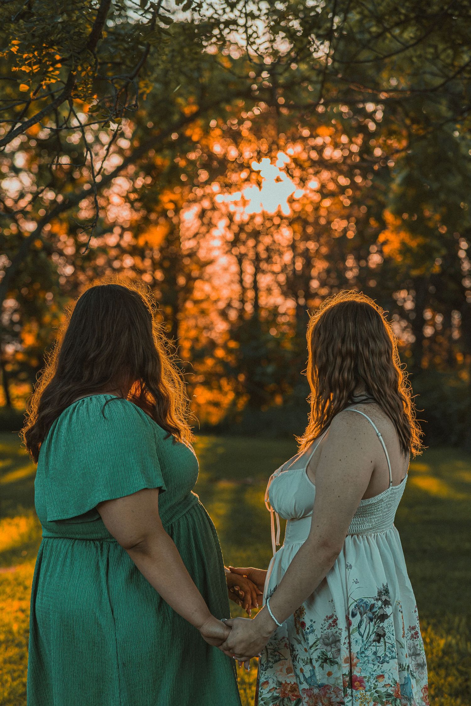 Two women in summer dresses hold hands facing a golden sunset through trees in a lush green park.