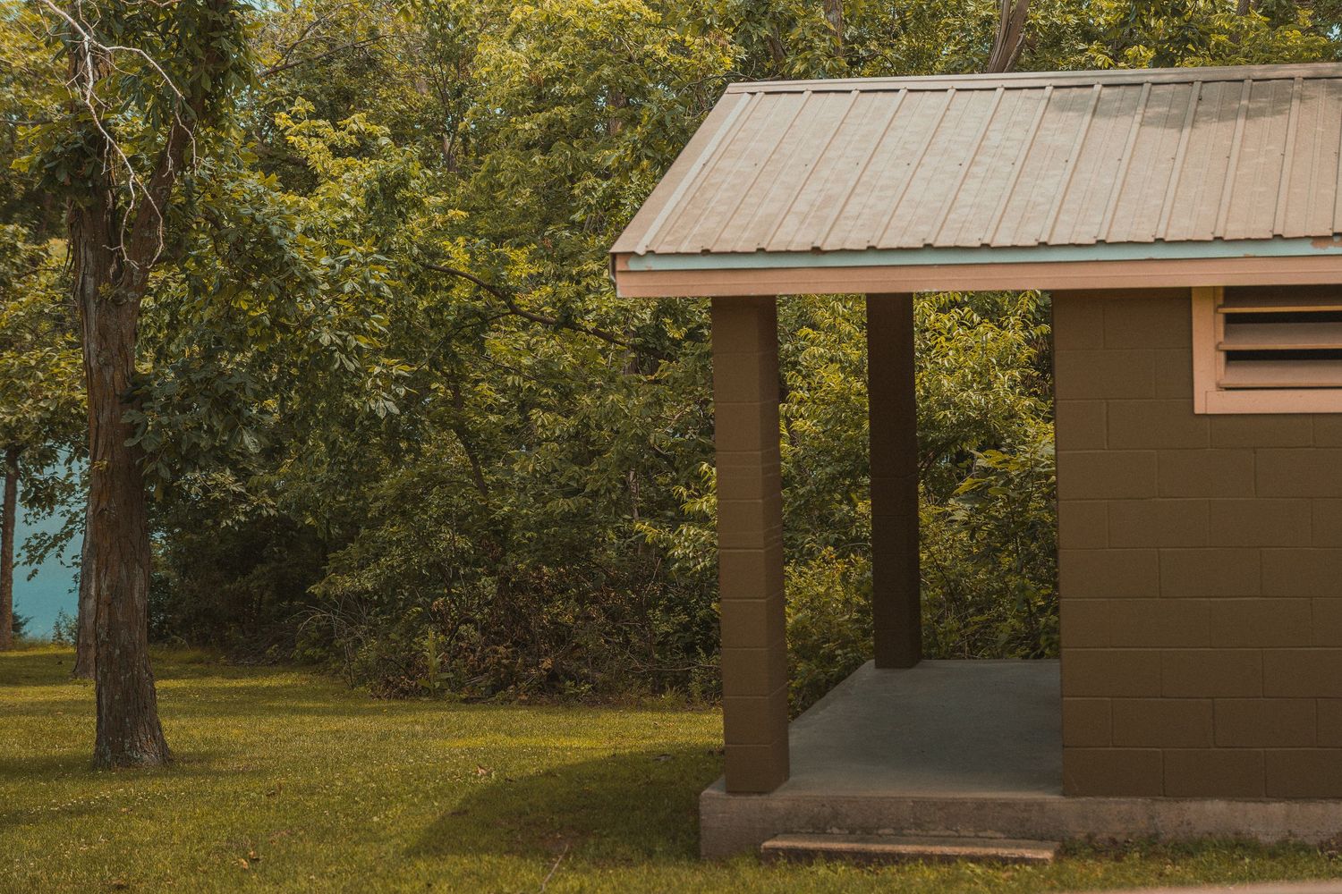 Small brown park shelter with metal roof surrounded by lush green trees on a grassy lawn.