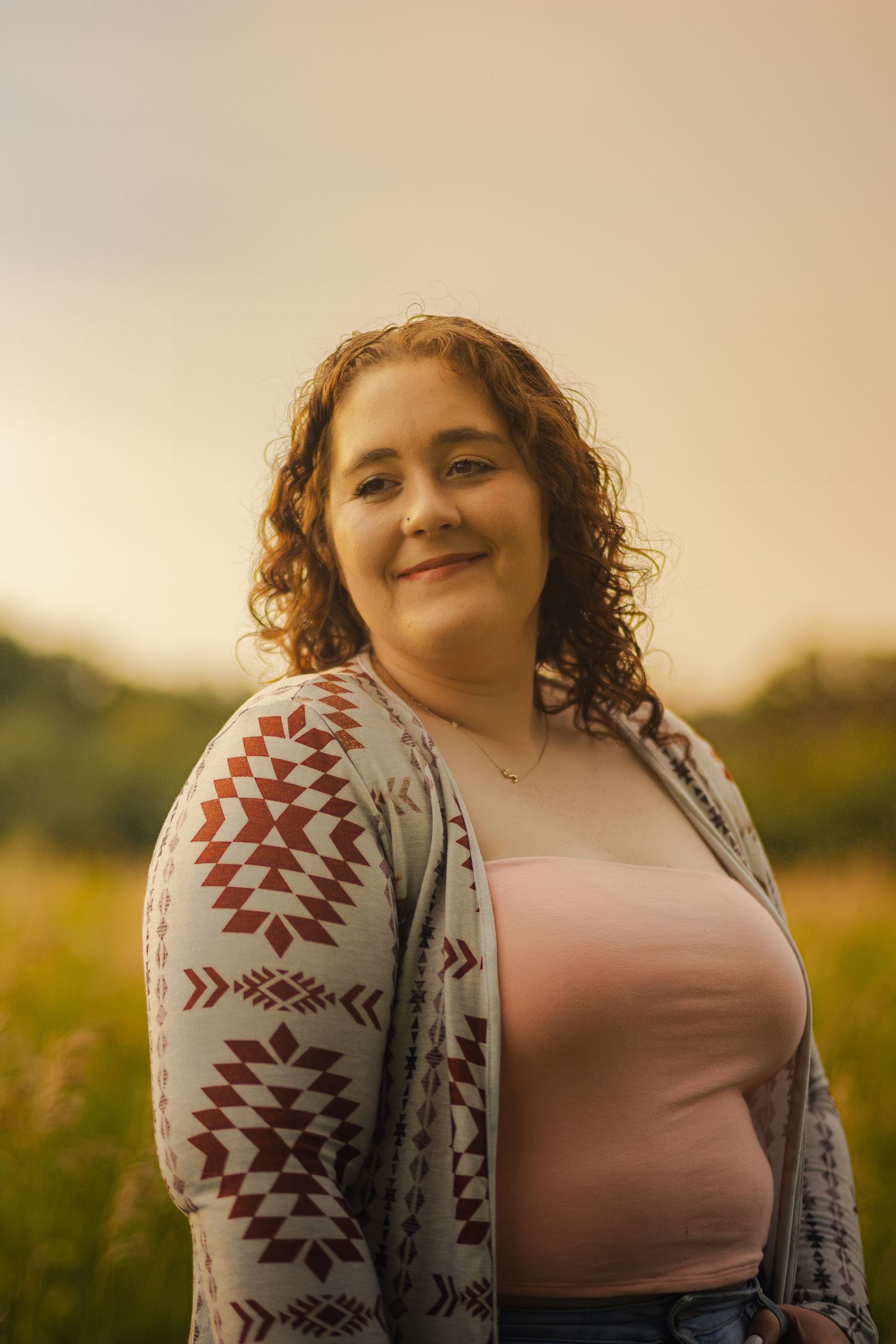 Young woman with curly hair smiling in a sunlit field, wearing a pink top and patterned cardigan at golden hour.