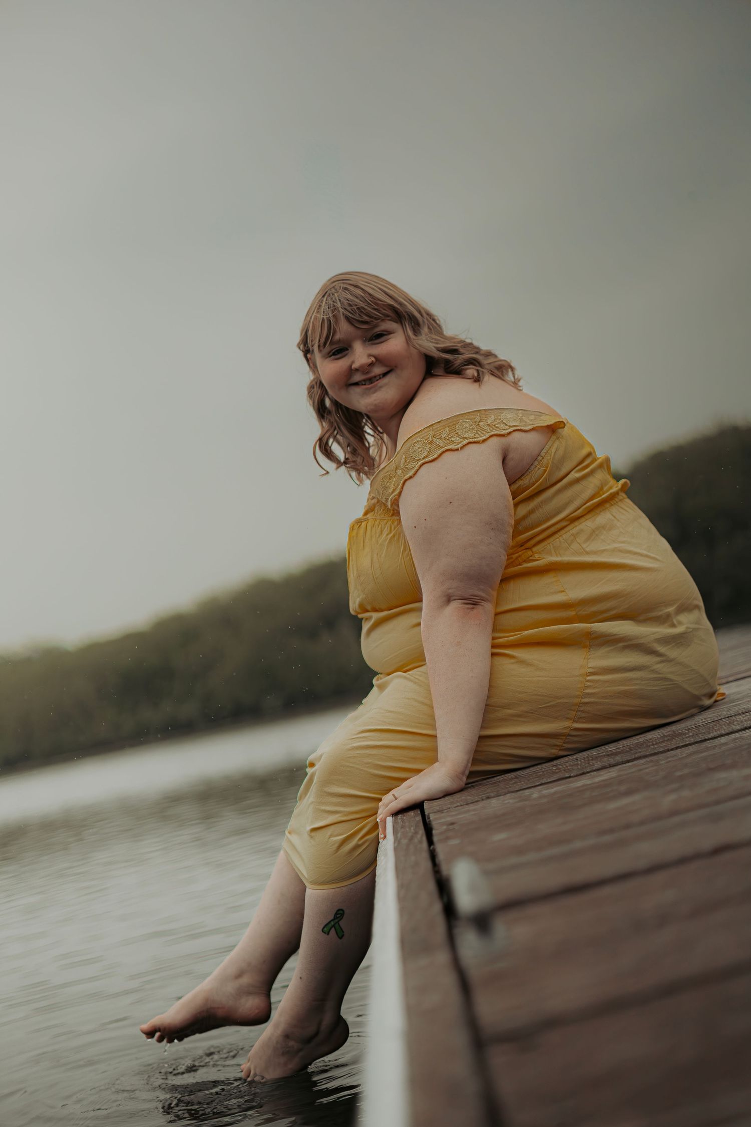 Smiling plus-size woman in yellow dress sitting on a wooden dock with feet in water, lake and trees in background.
