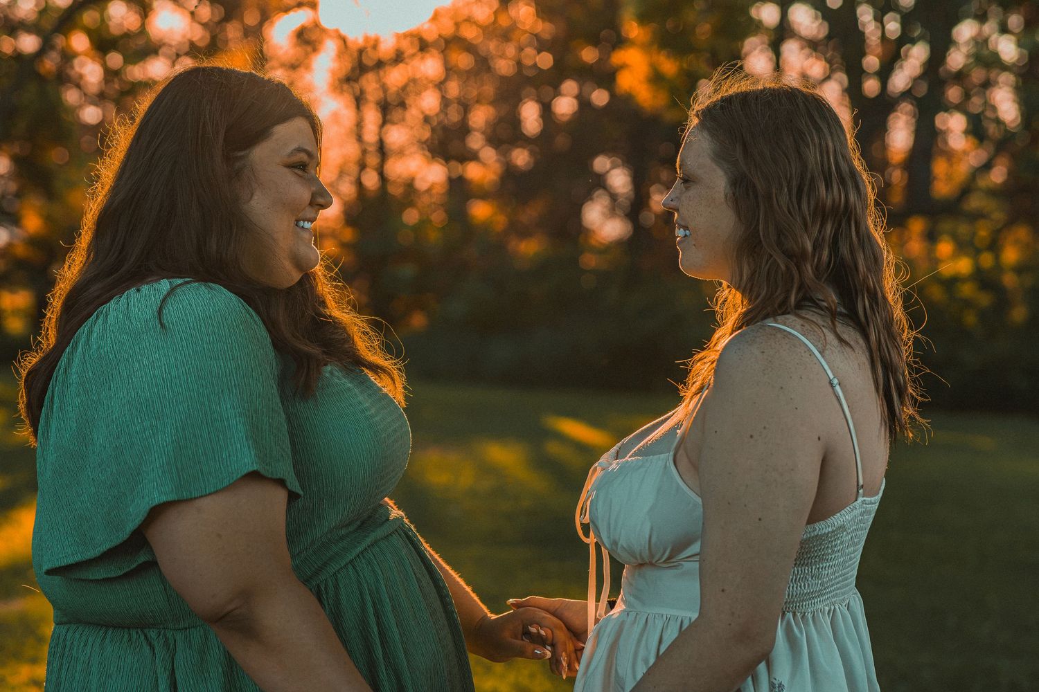Two women in green and white dresses hold hands at golden hour sunset in a lush outdoor setting.