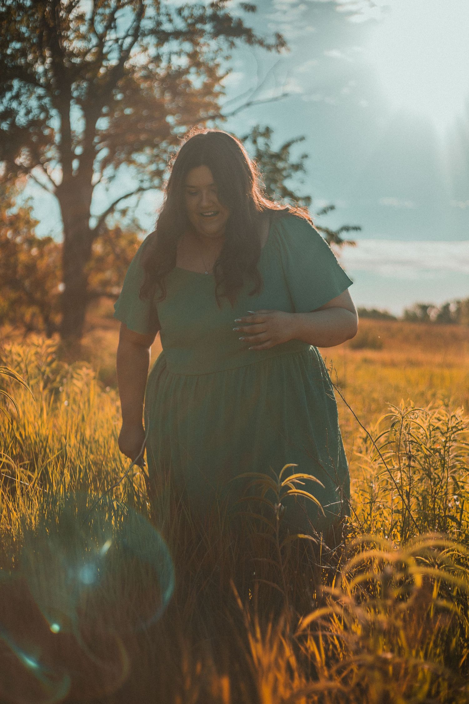 Pregnant woman in teal dress standing in golden field at sunset with warm lens flare and trees in background.