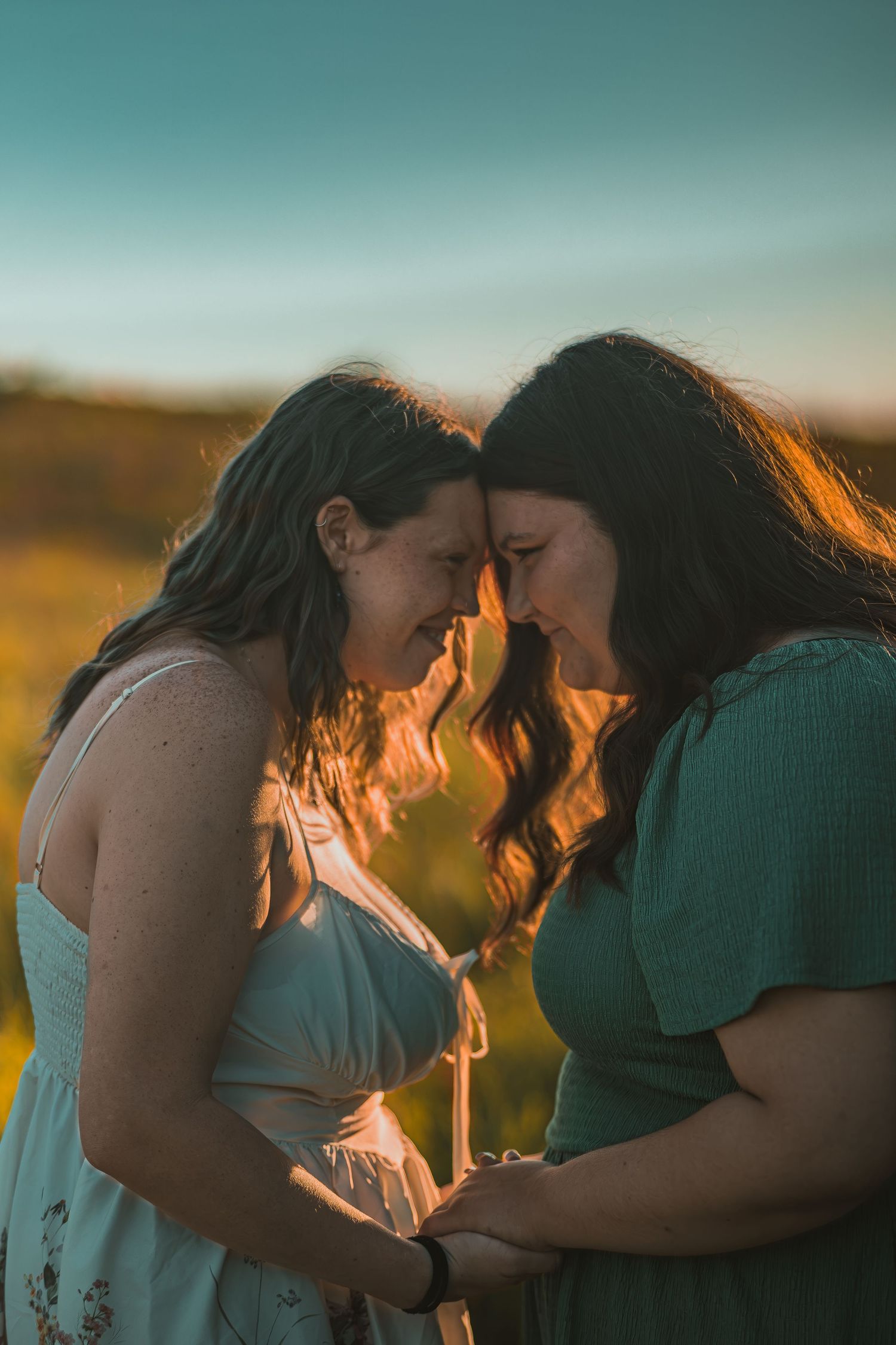 Two women touch foreheads tenderly at golden hour in a field, holding hands in a romantic outdoor portrait.