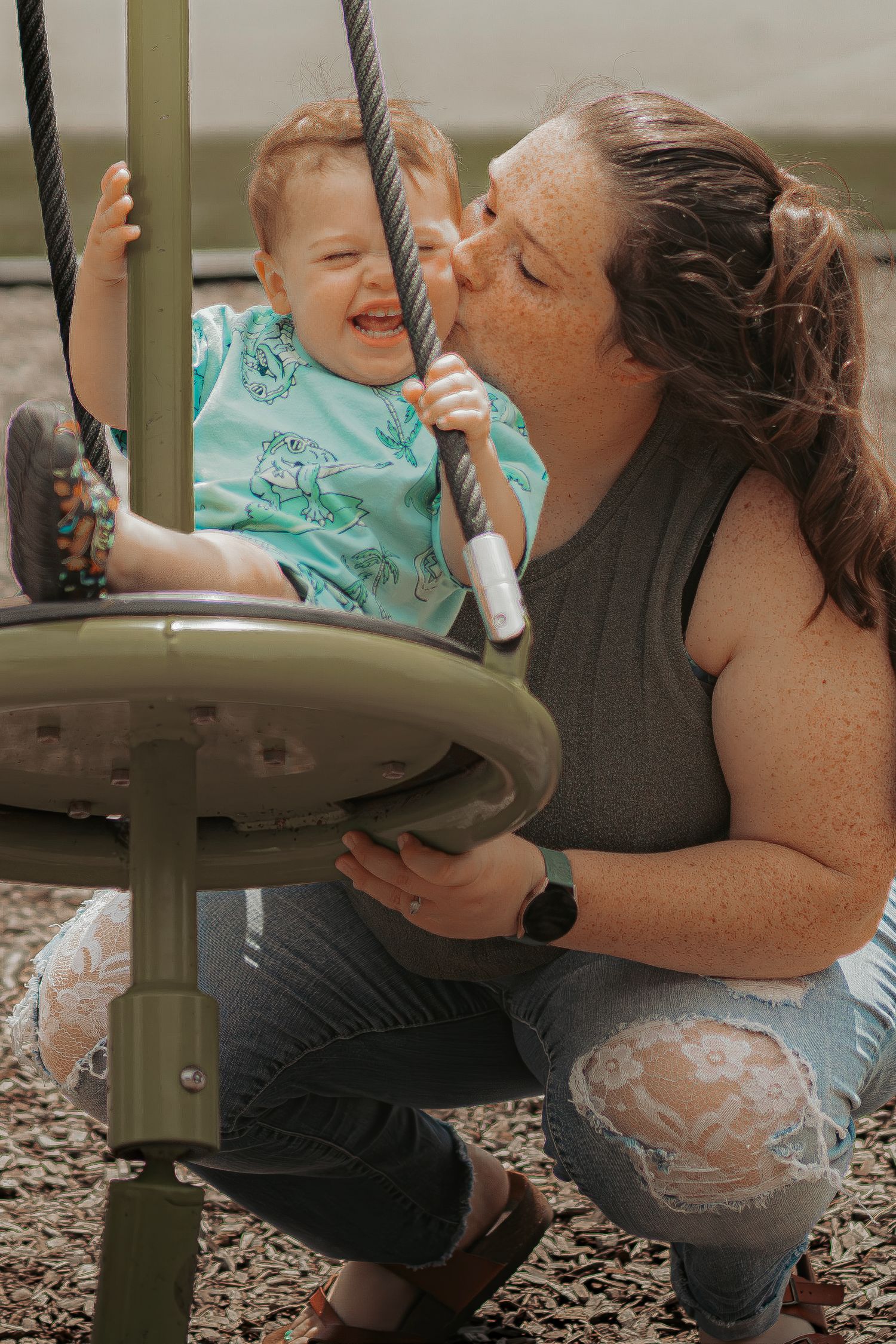 A mother kisses her laughing toddler sitting in a playground swing on a sunny day.