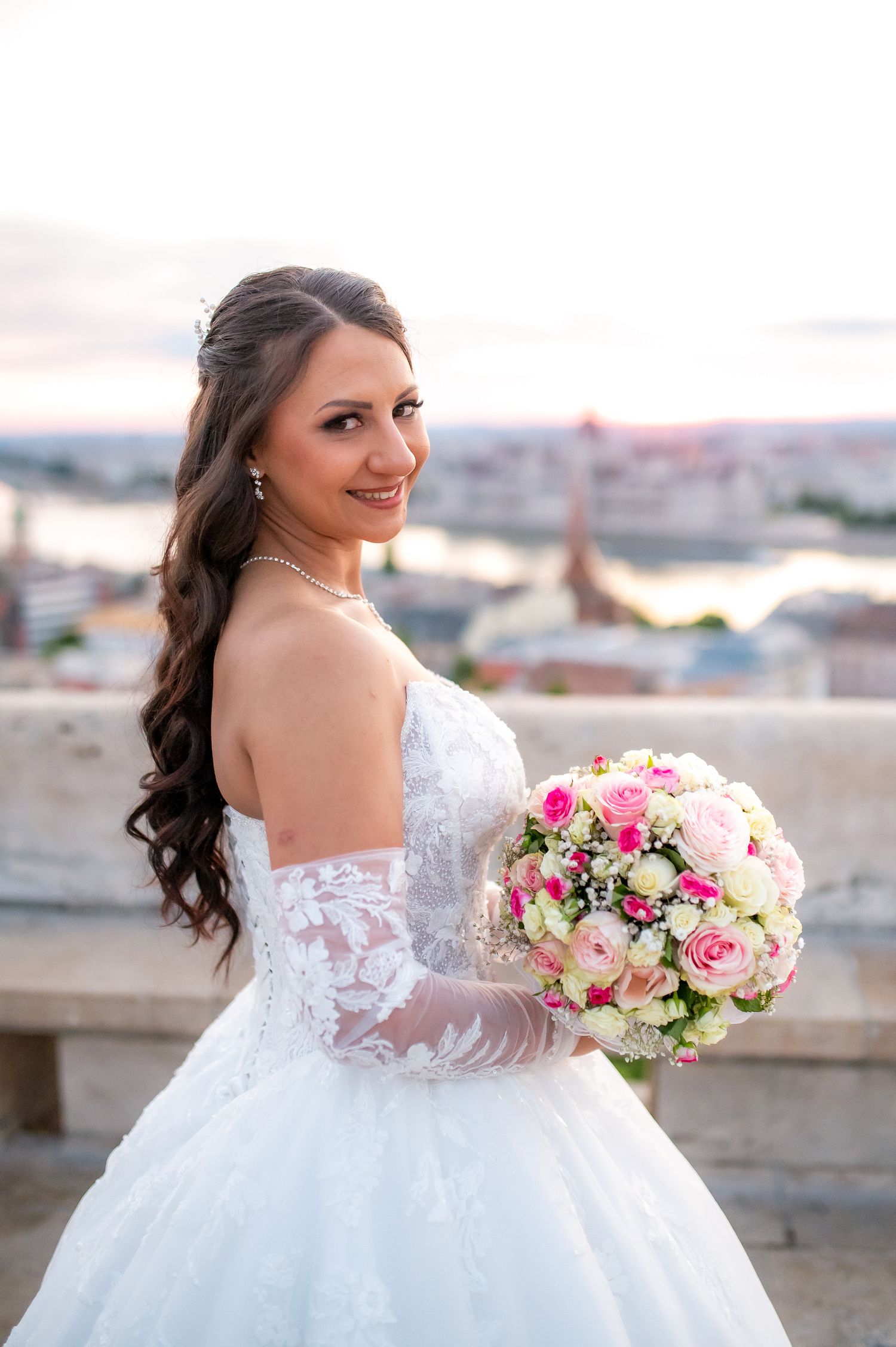 Smiling bride in white lace gown holds pink rose bouquet with city skyline backdrop at sunset.