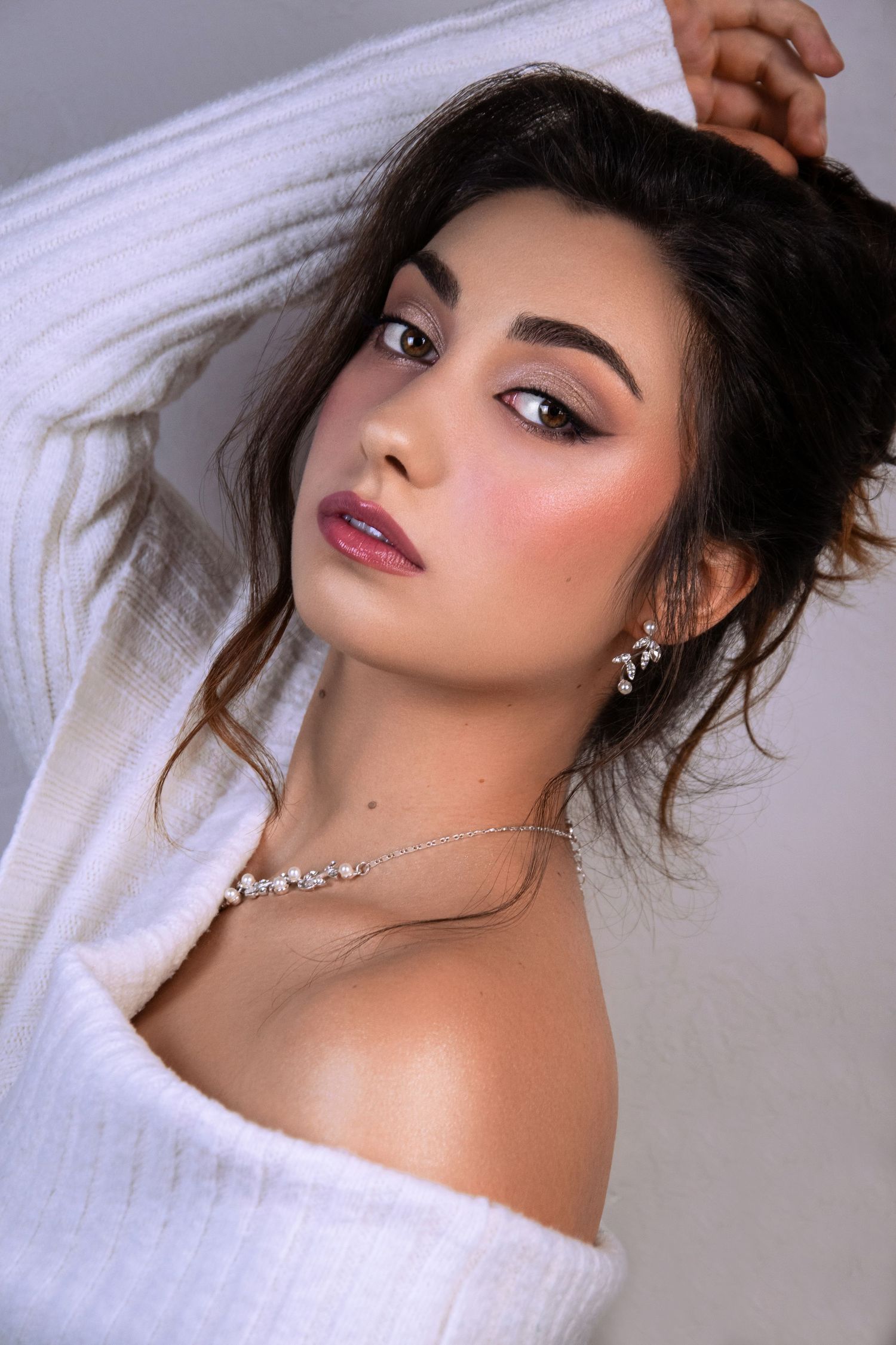 Young woman with smoky eye makeup, rosy blush, wearing white off-shoulder sweater and delicate pearl necklace and earrings.