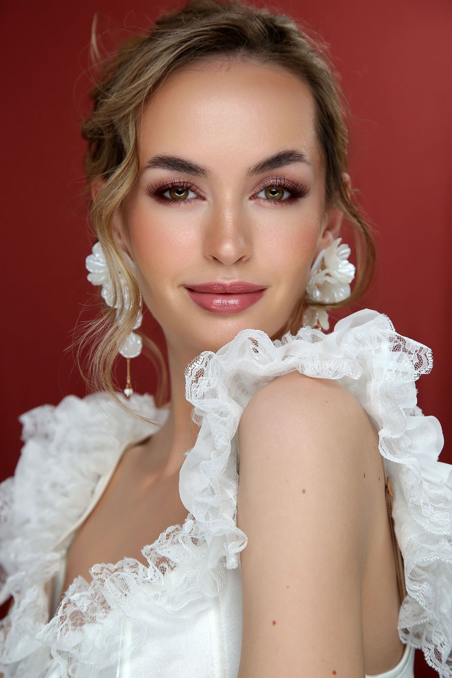 Young woman in white ruffled dress with floral earrings poses against a red background with elegant bridal makeup.