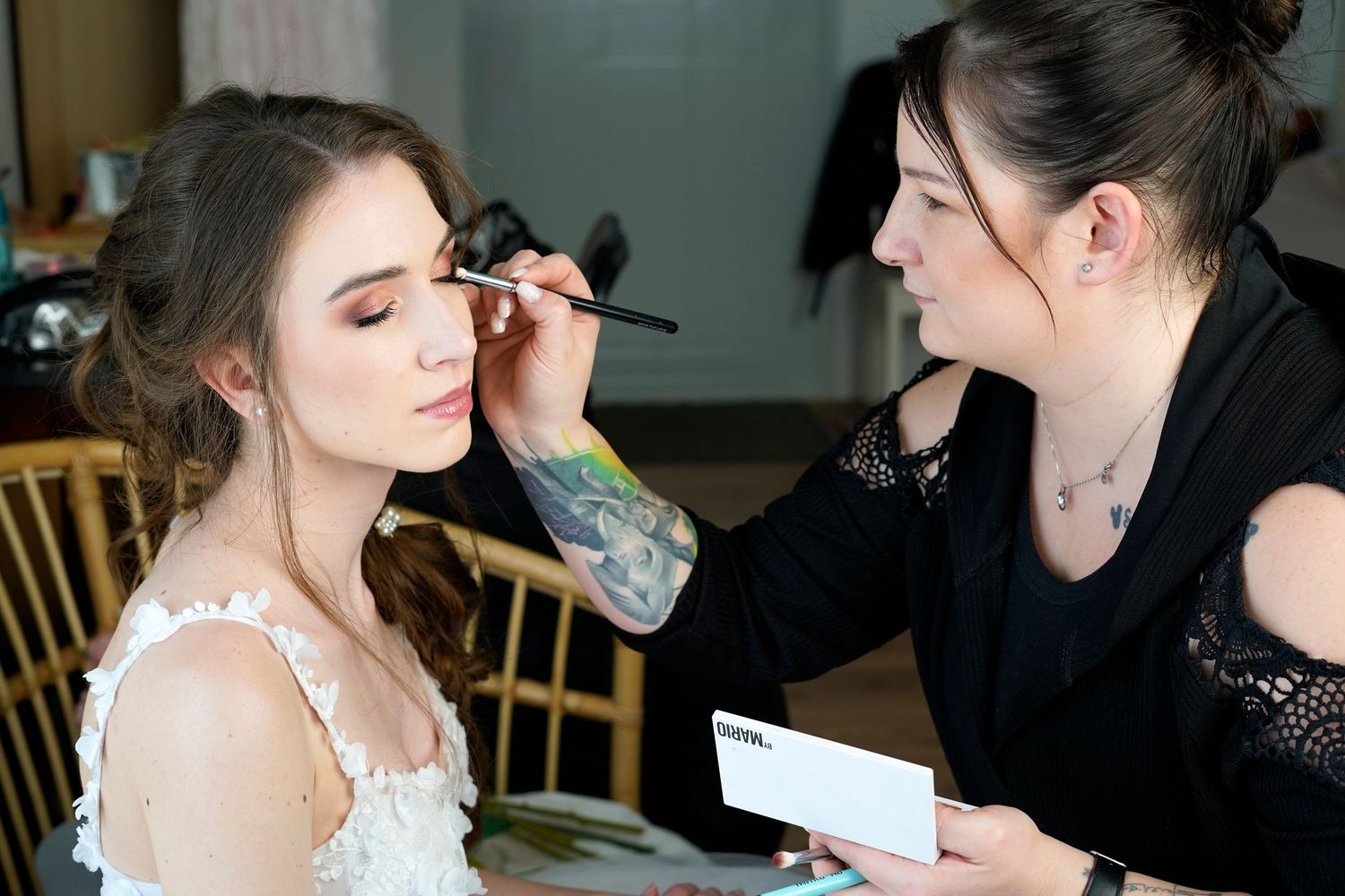A makeup artist applies eyeshadow to a bride wearing a white lace dress during wedding preparations.