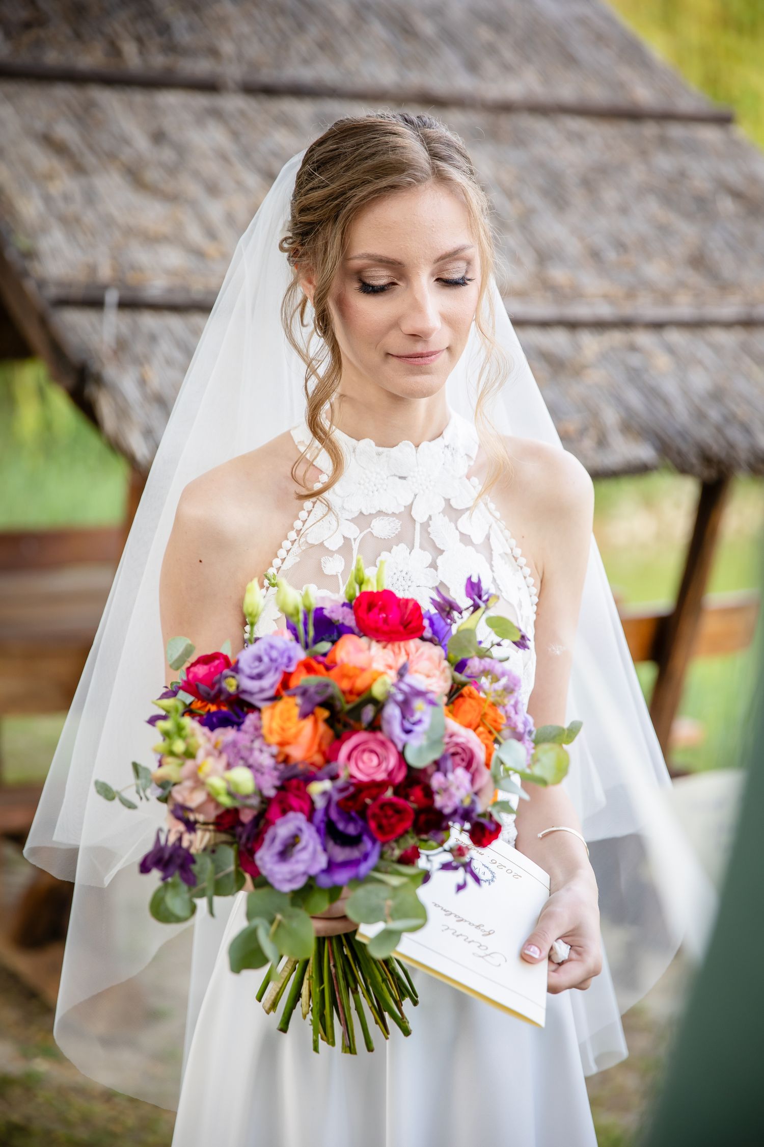 Bride in white lace dress and veil holds colorful bouquet of red, purple, and orange flowers outdoors.