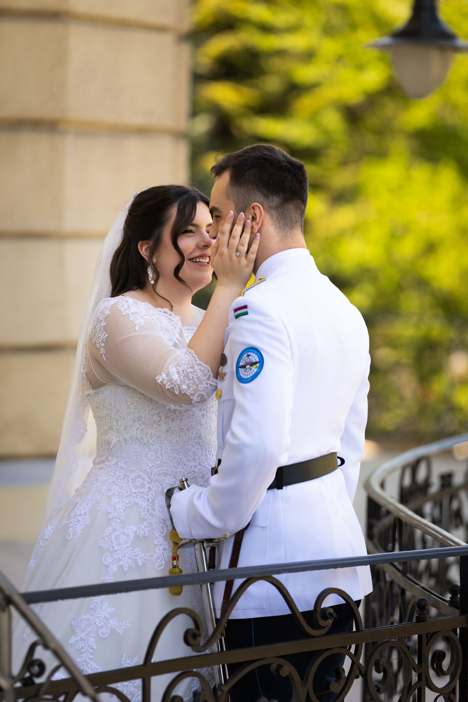 A bride in white lace gown and groom in military uniform share a romantic moment on a balcony.