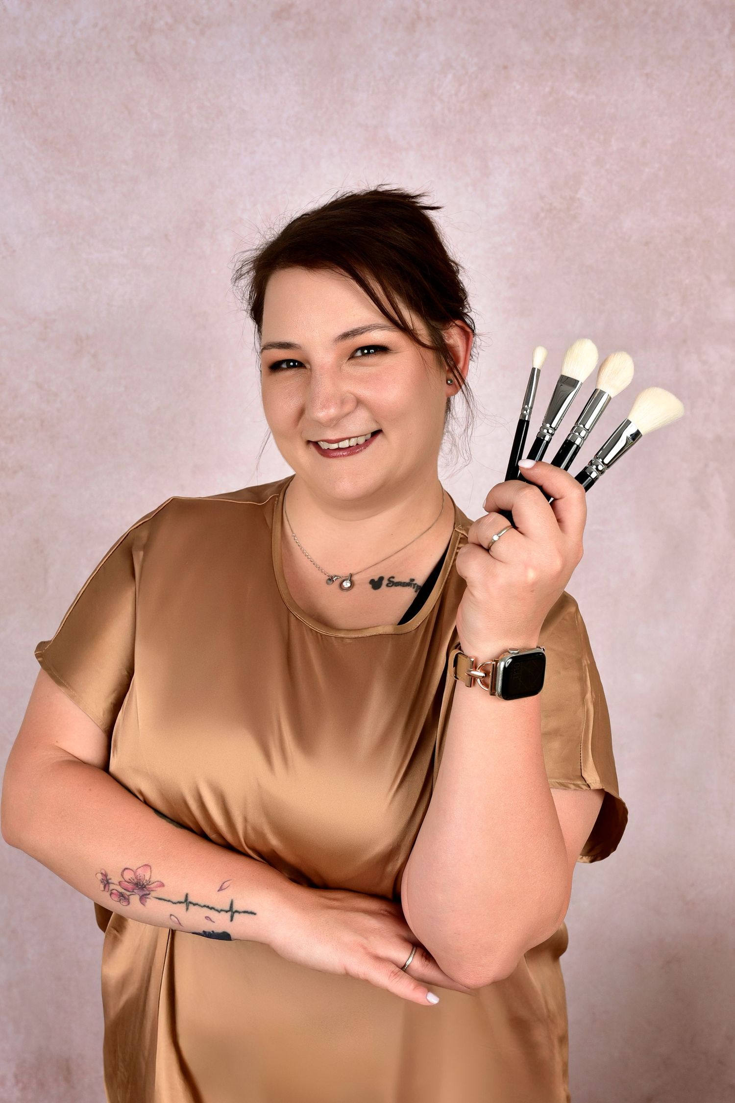 Professional makeup artist in tan blouse holding brushes while showing arm tattoo against neutral studio backdrop.
