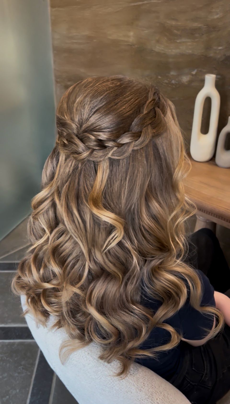 Half-up braided crown hairstyle with loose curls and blonde highlights on long brown hair.