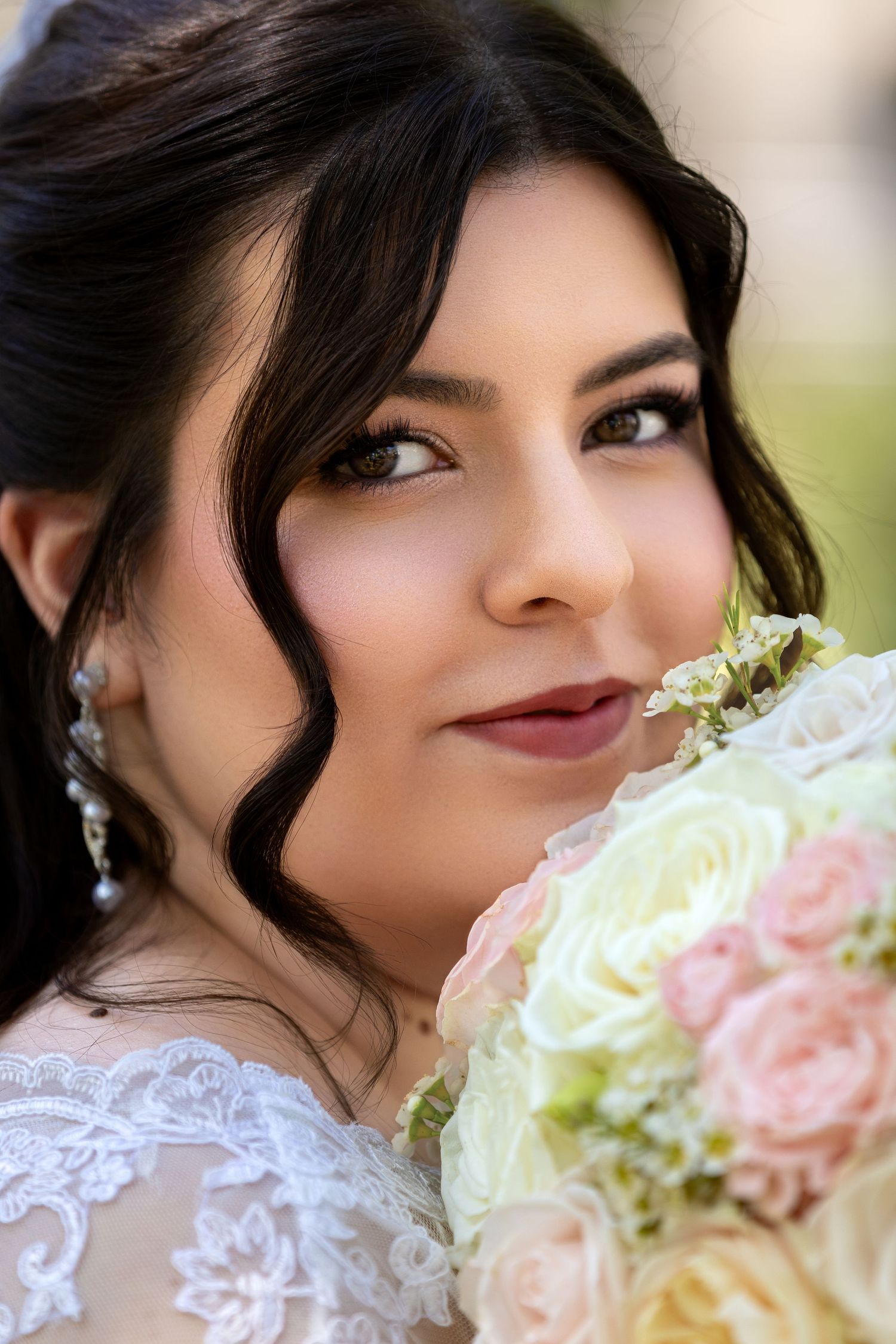 A beautiful bride with dark hair holds a bouquet of pink and white roses, wearing lace dress and pearl earrings.