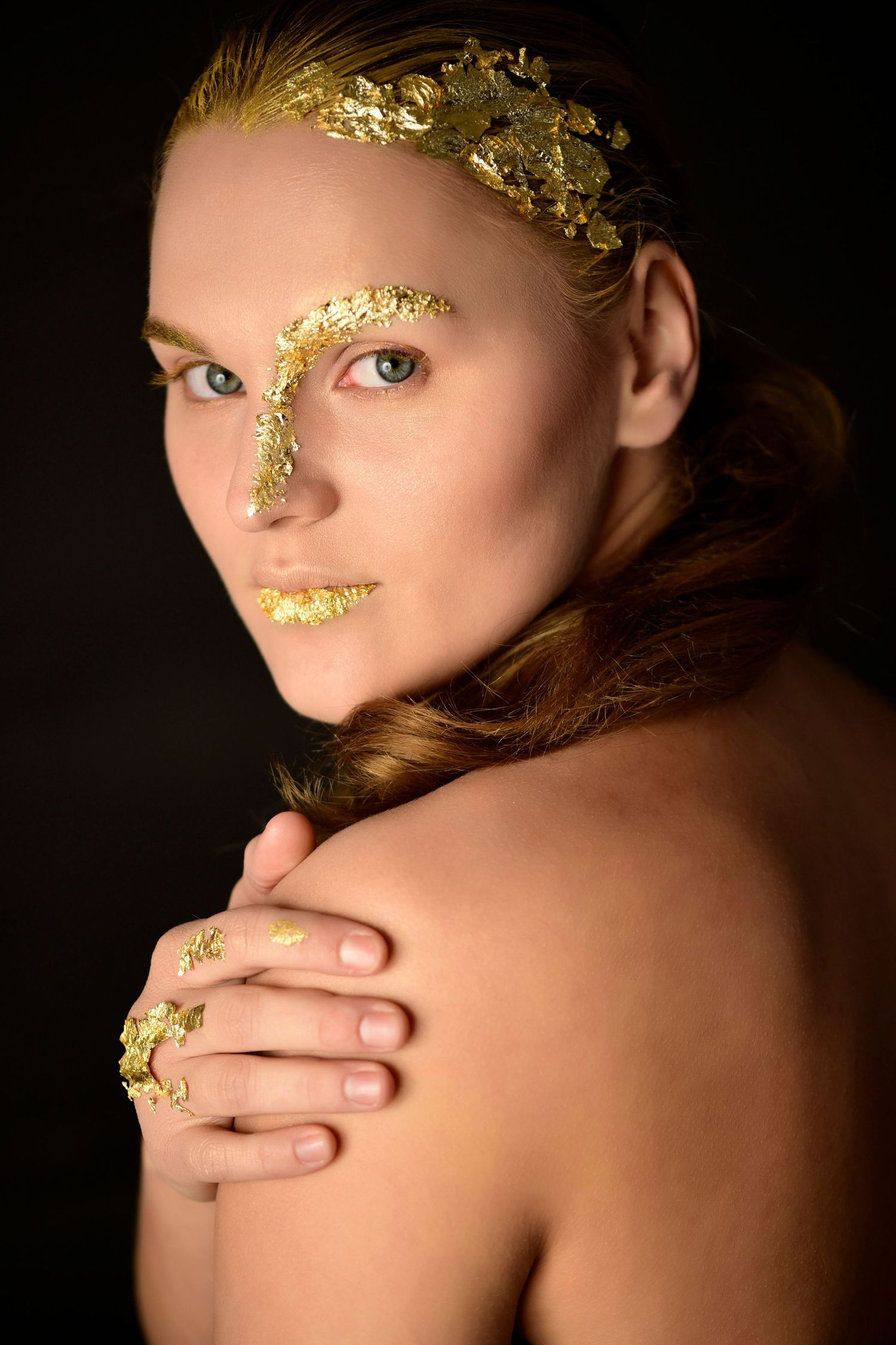 Creative beauty portrait featuring gold leaf makeup and bare shoulders against dark background.