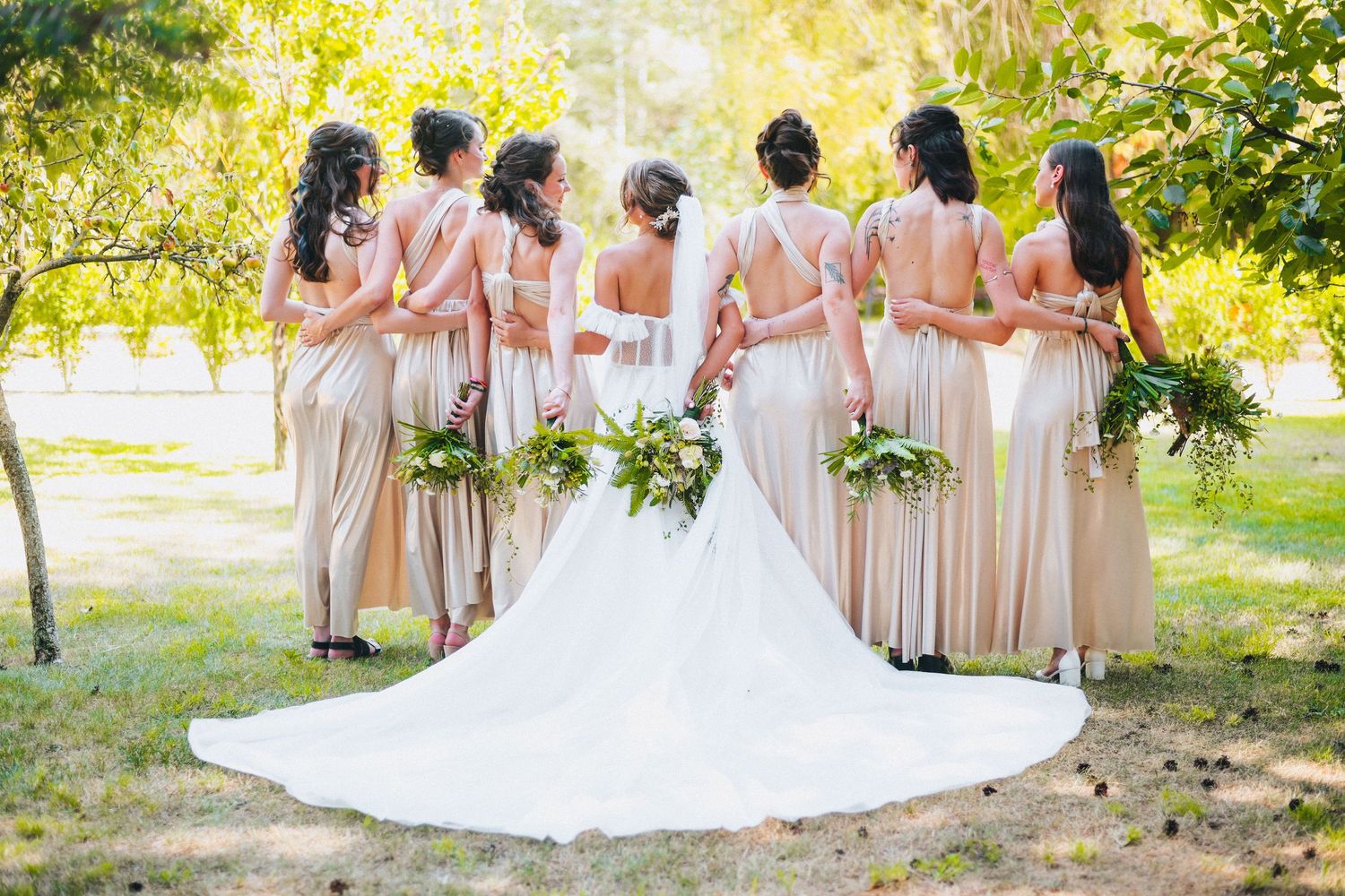 Bride and bridesmaids in champagne gowns holding greenery bouquets, posing with backs to camera in lush garden.