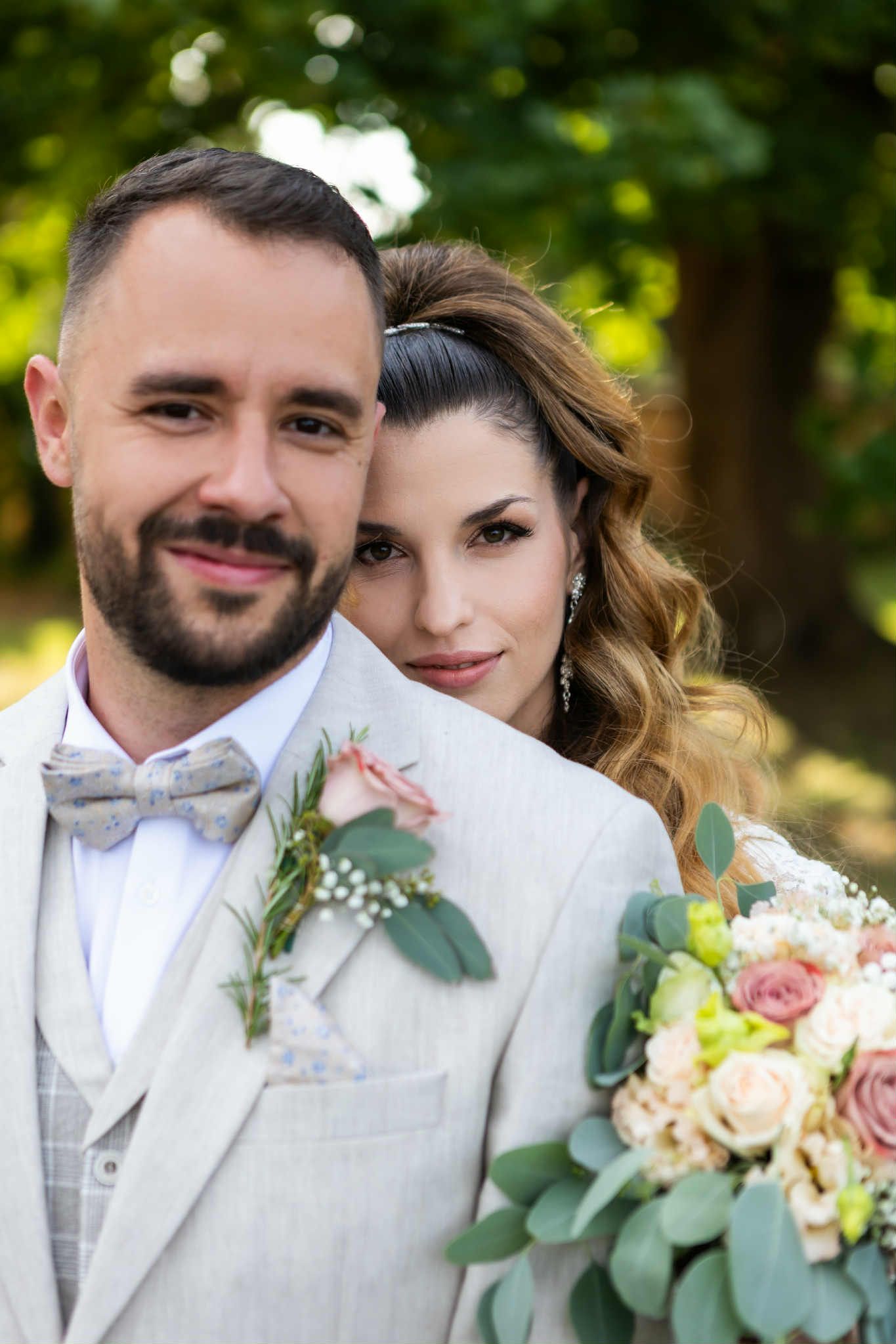 A couple poses on their wedding day with a beautiful bouquet of roses and eucalyptus against a blurred green background.