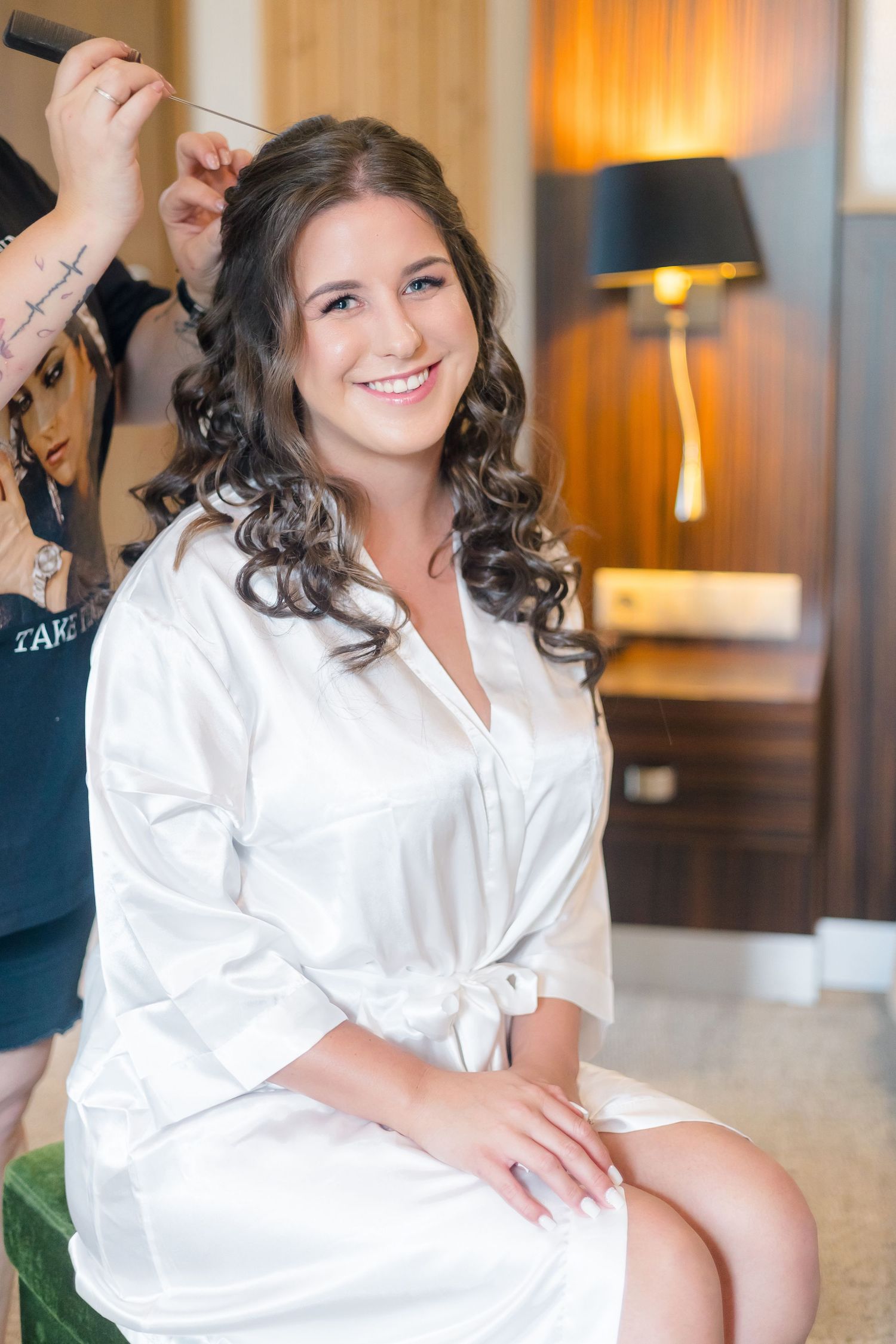 A person wearing a white silk robe sits in a chair while having their curled hair styled by a stylist in the background.