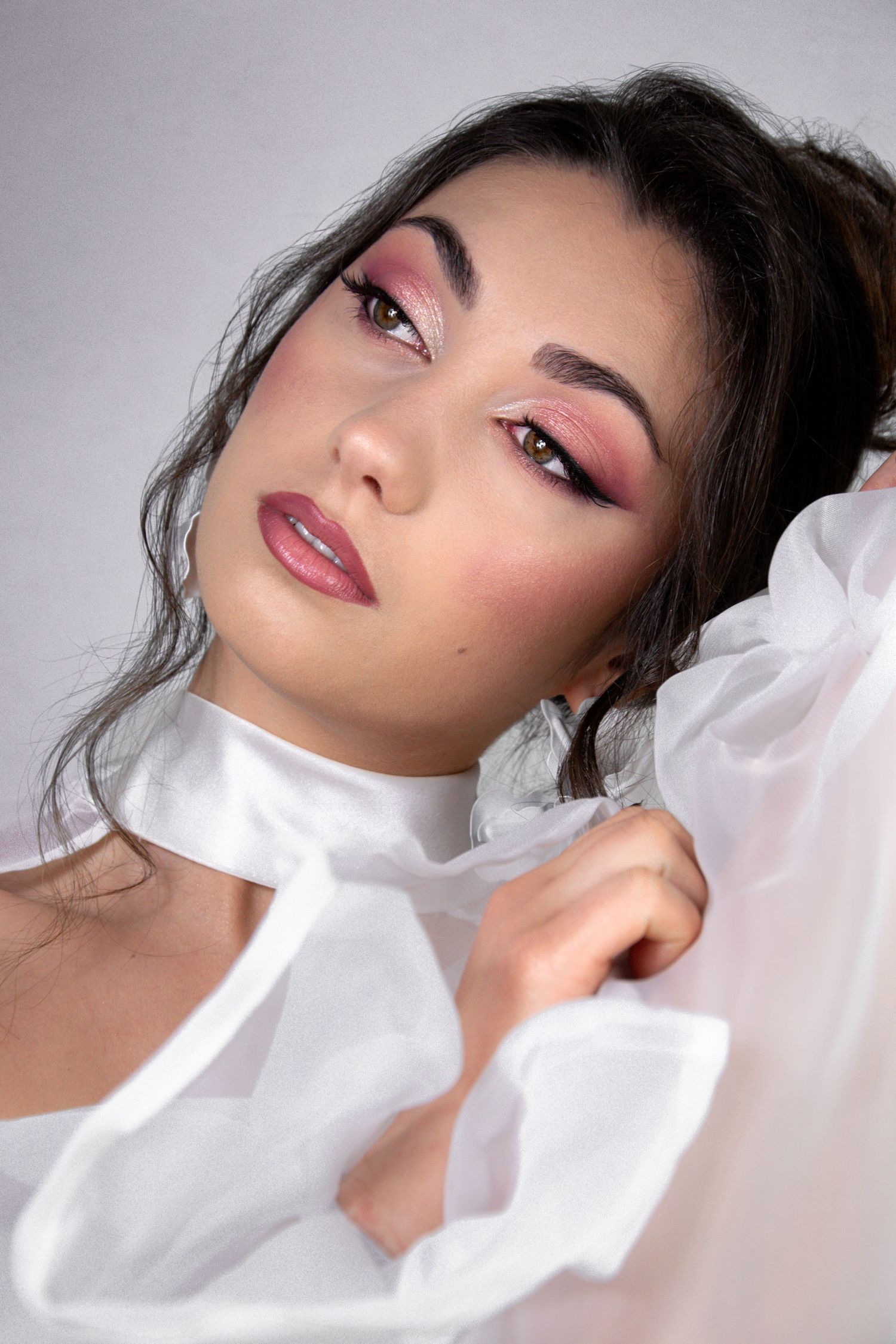 Young woman with pink eye makeup and glossy lips wearing a white ruffled blouse, posing gracefully.