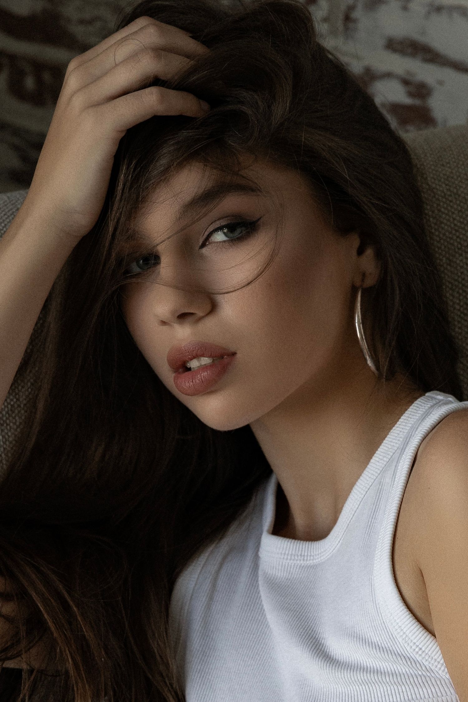 A moody portrait shows a person with long dark hair wearing a white tank top and hoop earrings in soft lighting.