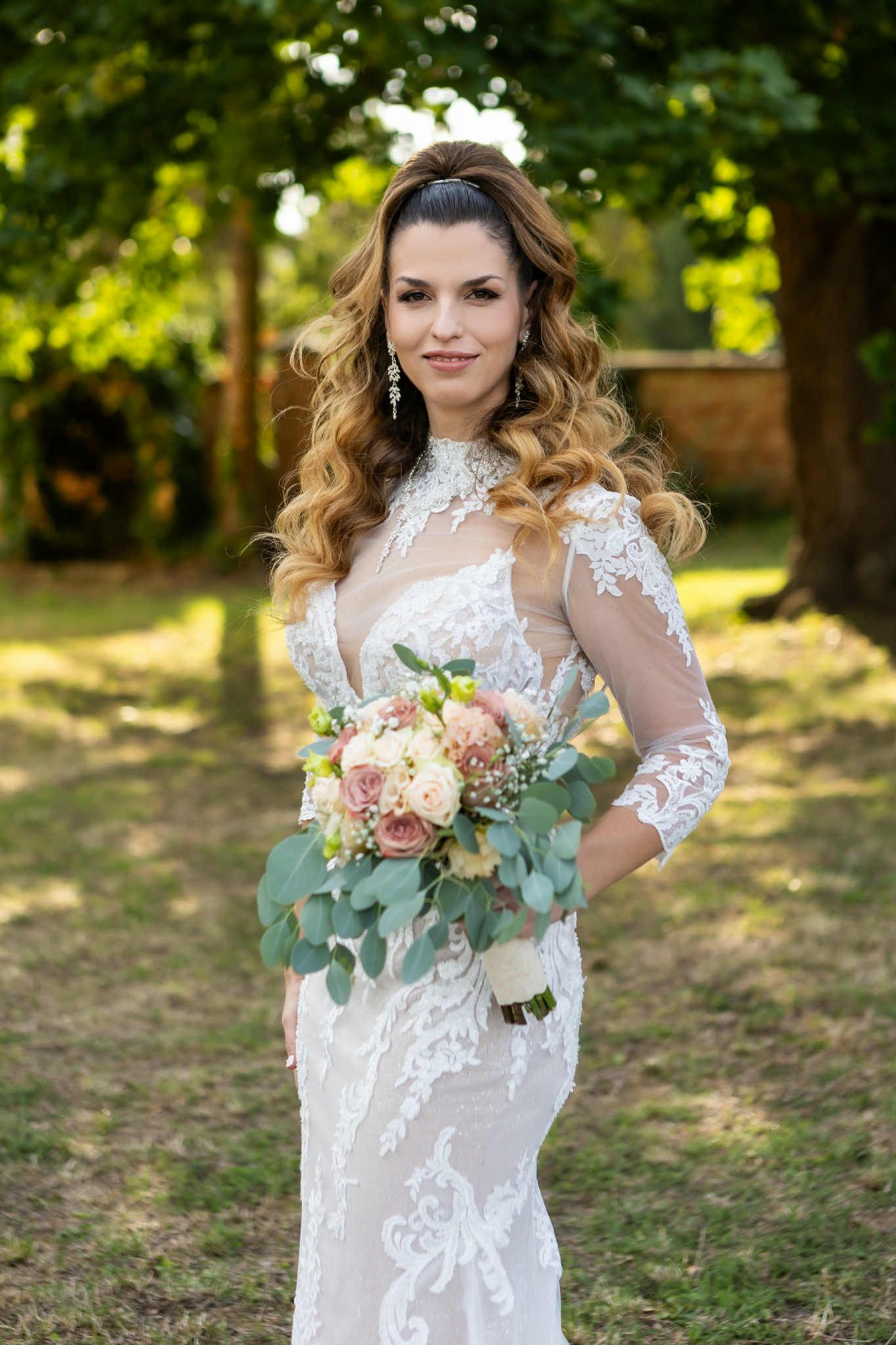Beautiful bride in lace white dress holds elegant bouquet with eucalyptus and roses in outdoor garden setting.