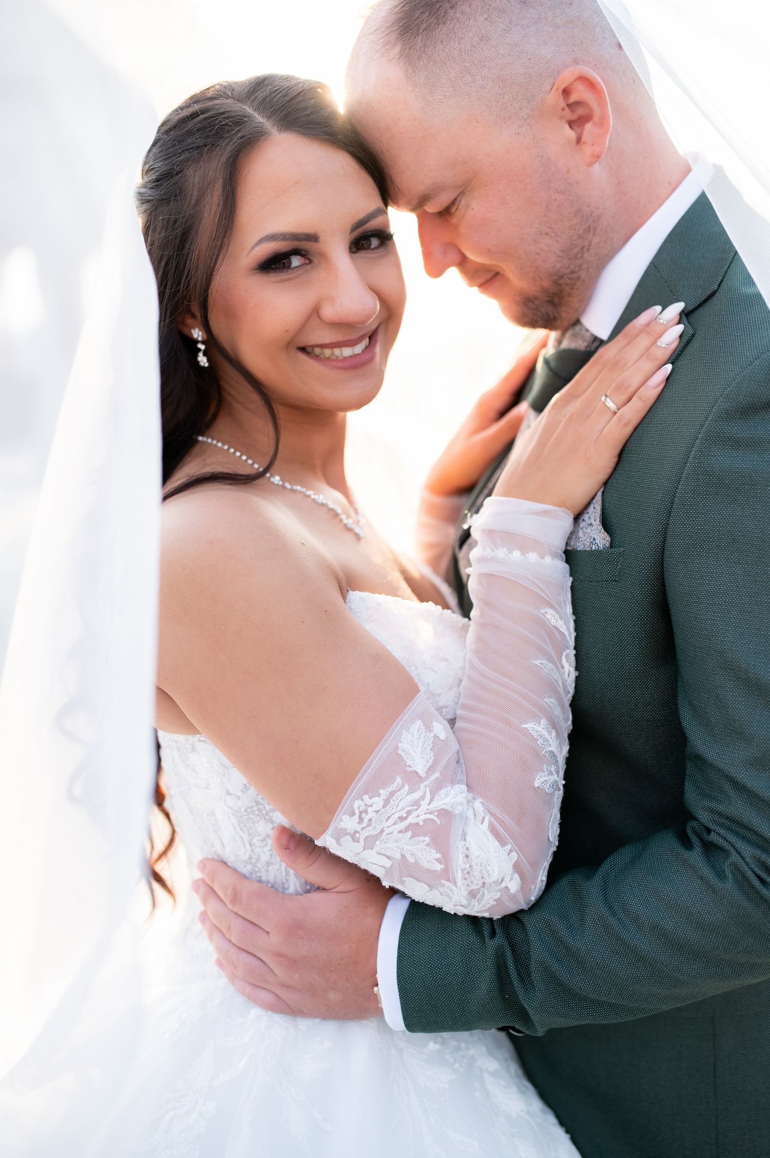 Smiling bride in lace gown embraces groom in green suit, foreheads touching in a romantic wedding portrait.