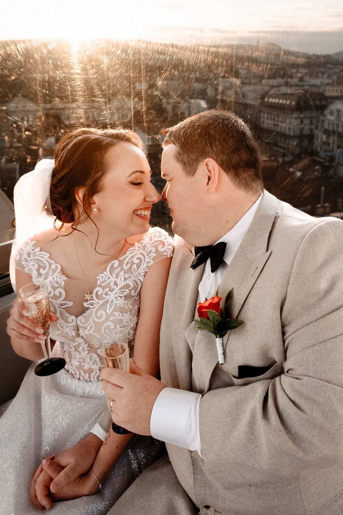 Bride and groom share a romantic moment with champagne at sunset, overlooking a city skyline on their wedding day.