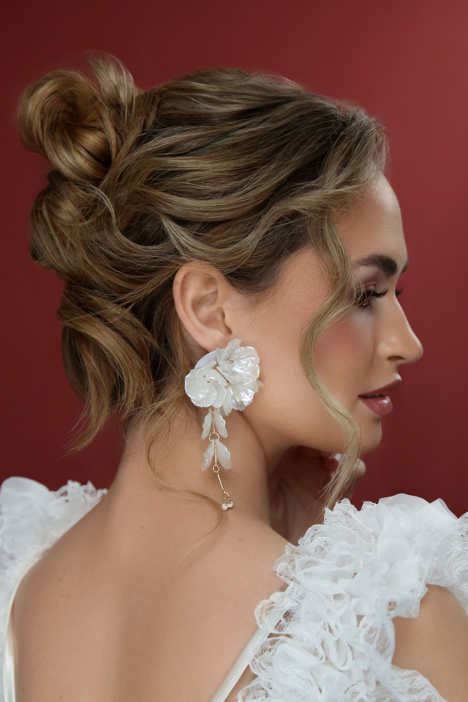 Bride with elegant updo hairstyle wearing large white floral drop earrings and ruffled white gown against red background.