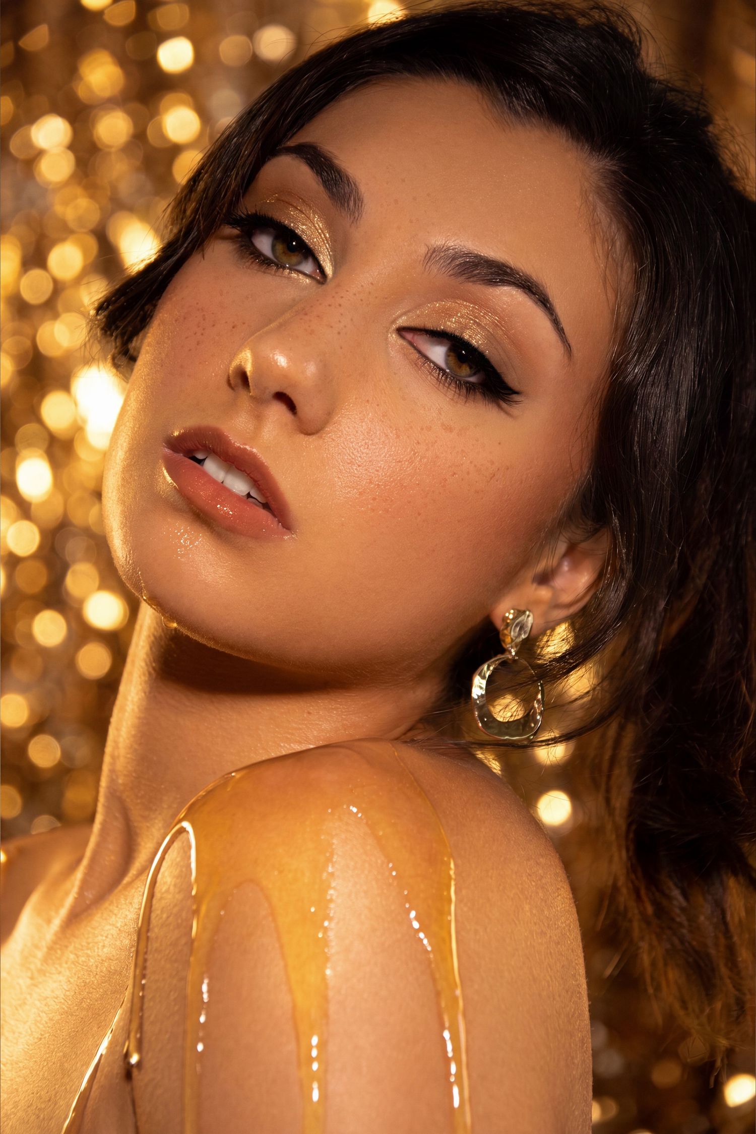 Woman with golden glitter makeup, winged eyeliner, and gold earrings, with honey dripping on her shoulder against bokeh background.