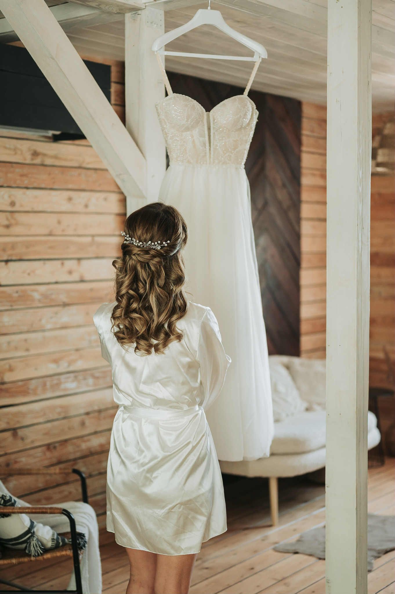 A white wedding dress hangs on display while someone in a silk robe with curled hair views it from behind.