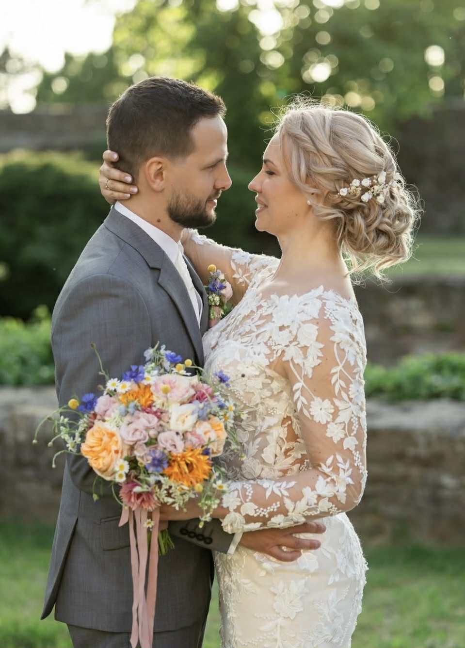Couple shares intimate moment in garden wedding setting with colorful peach and purple bouquet and lace dress details.