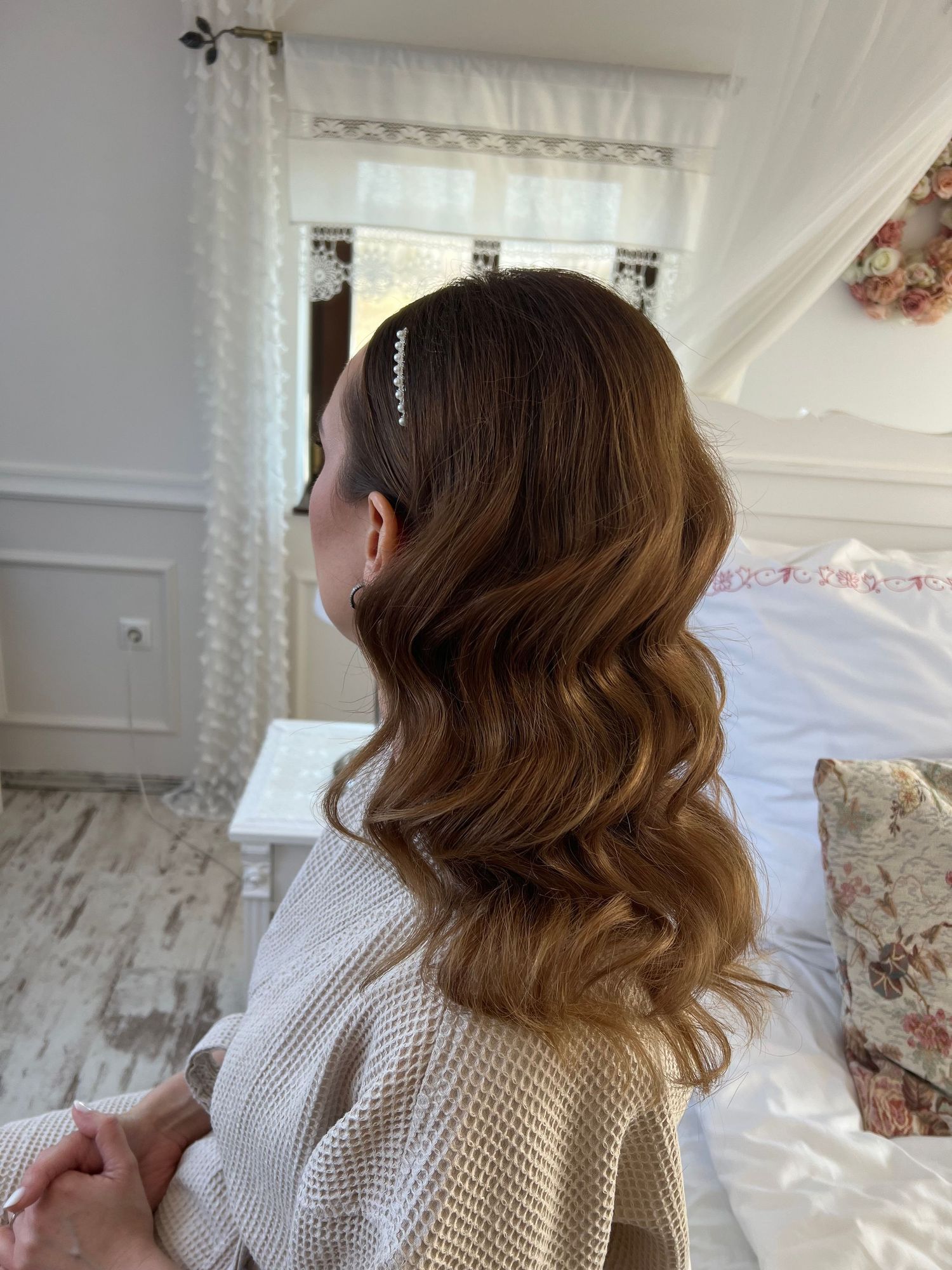 Woman with long wavy auburn hair styled with a pearl headband, sitting in a bright white shabby chic bedroom.