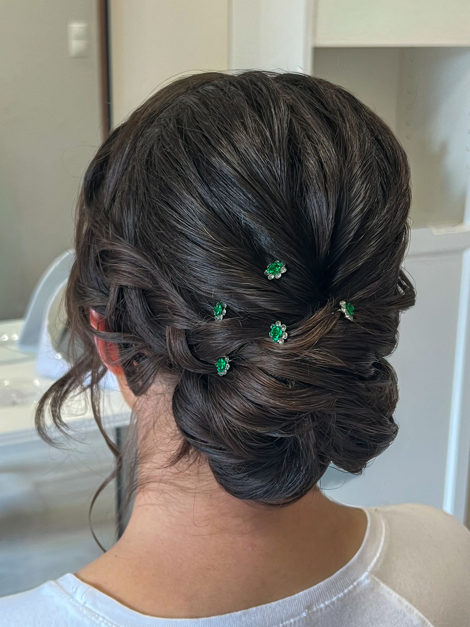 Elegant dark hair updo with braided detail and small green floral hair pins, styled for a formal occasion.