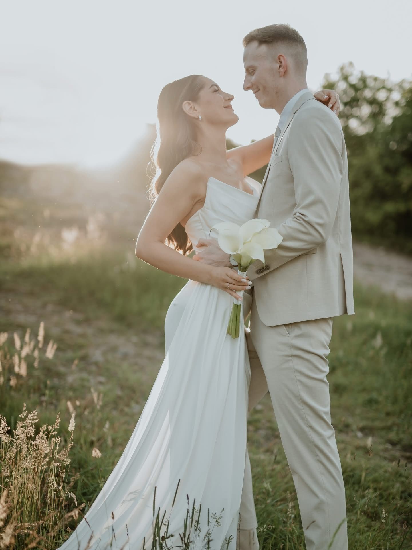 Romantic couple poses in a sunlit meadow, dressed in white and beige, during an outdoor wedding photo session.