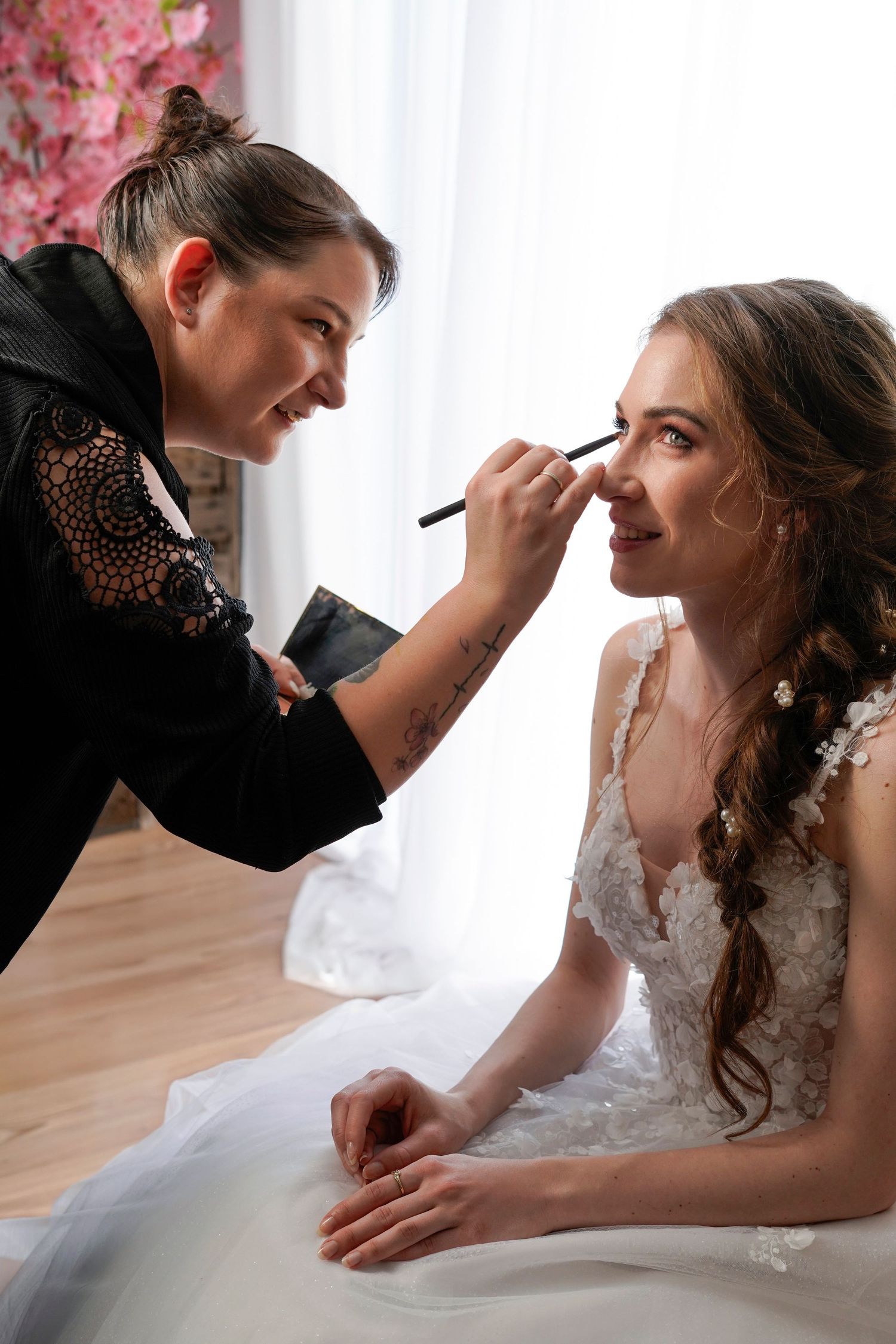 A makeup artist applies eye makeup on a bride wearing a white lace wedding dress beside a window with pink florals.