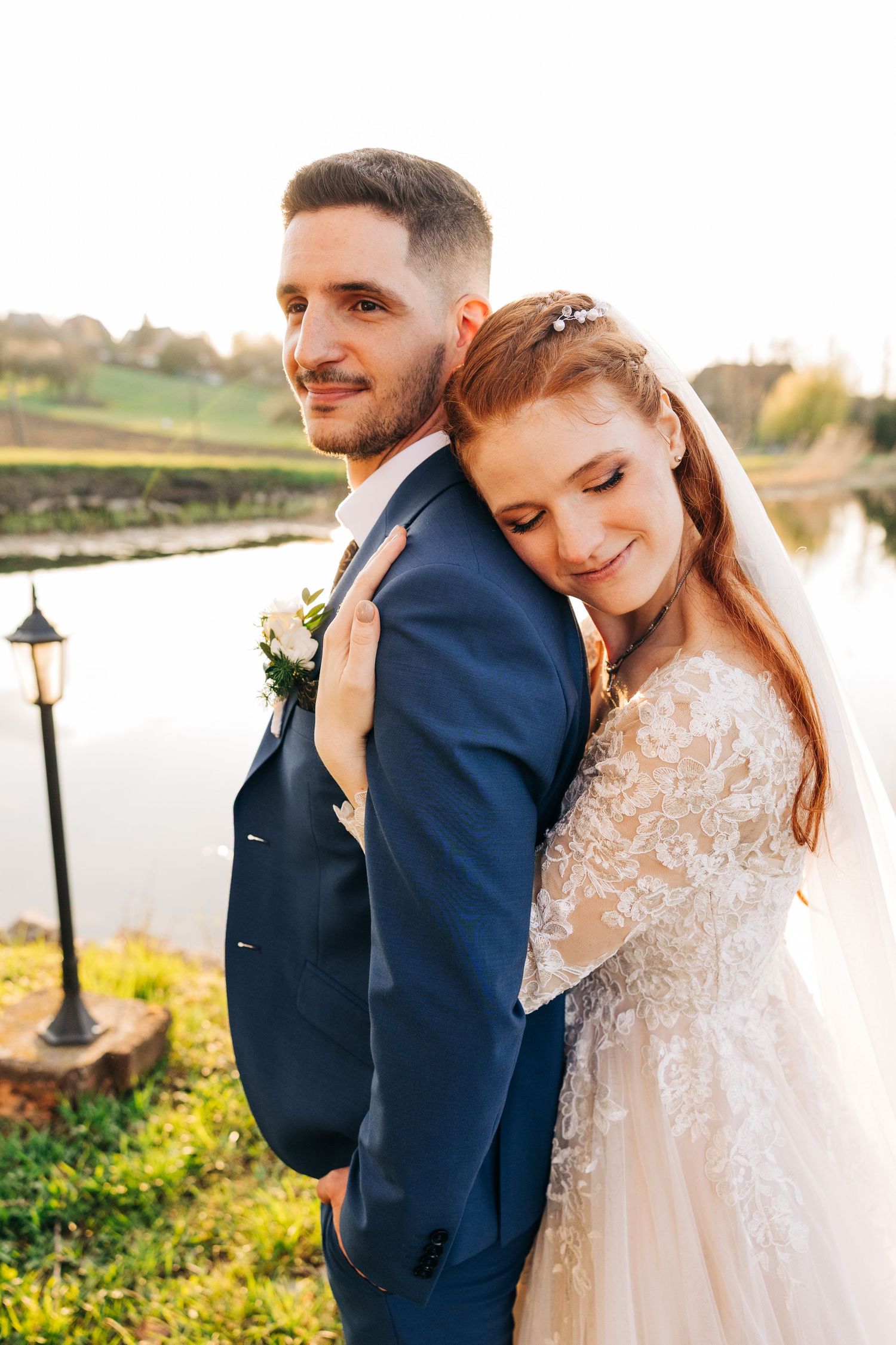 A couple in wedding attire embrace by a lakeside setting with rustic lampposts and golden sunlit landscaping.