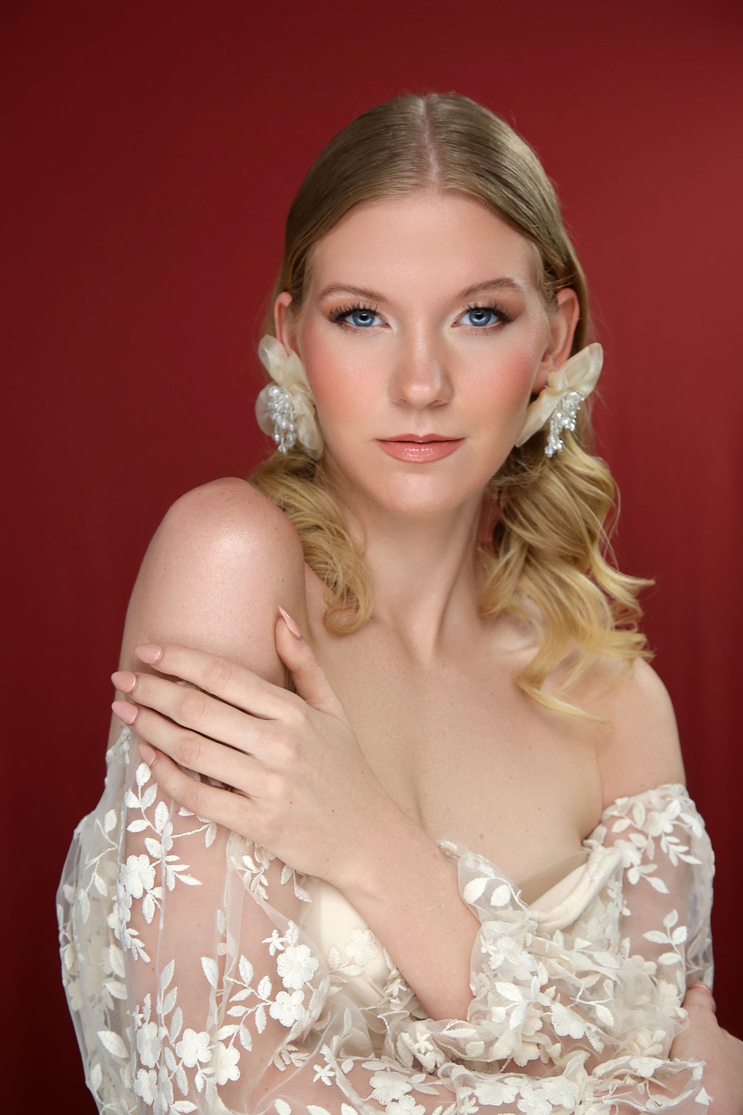 Blonde woman in white floral lace gown with pearl earrings poses against red background.