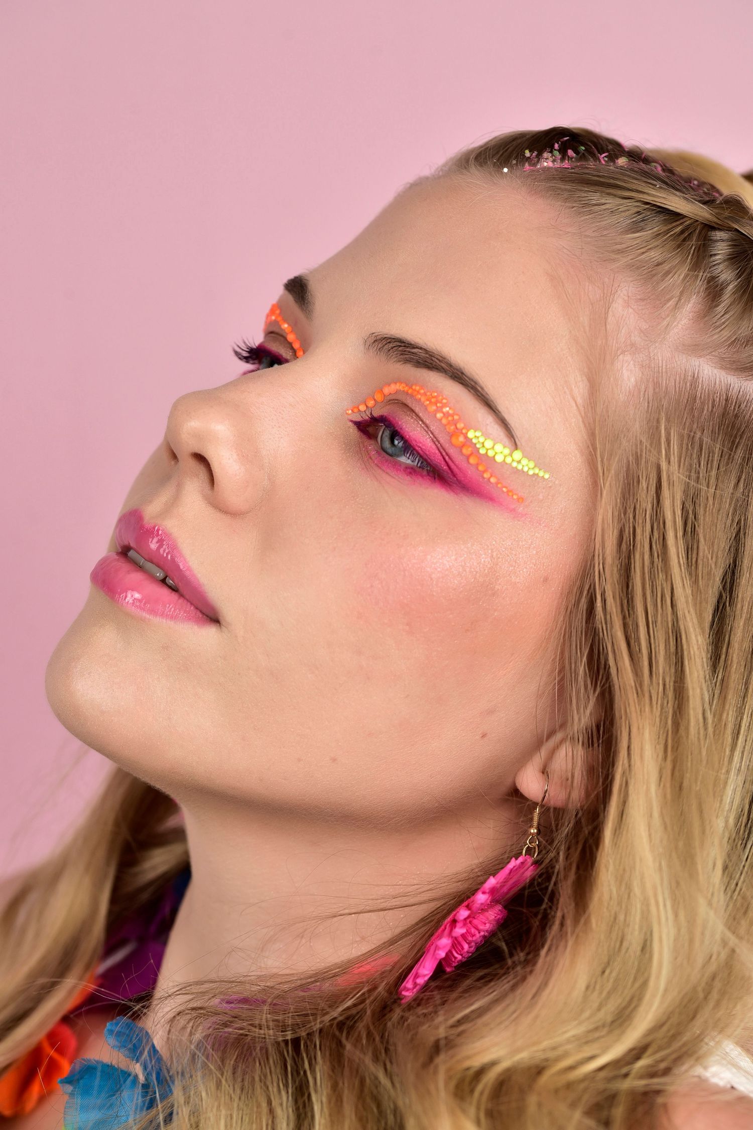 Close up beauty shot with vibrant pink and orange eye makeup and gold accent against a pink background.