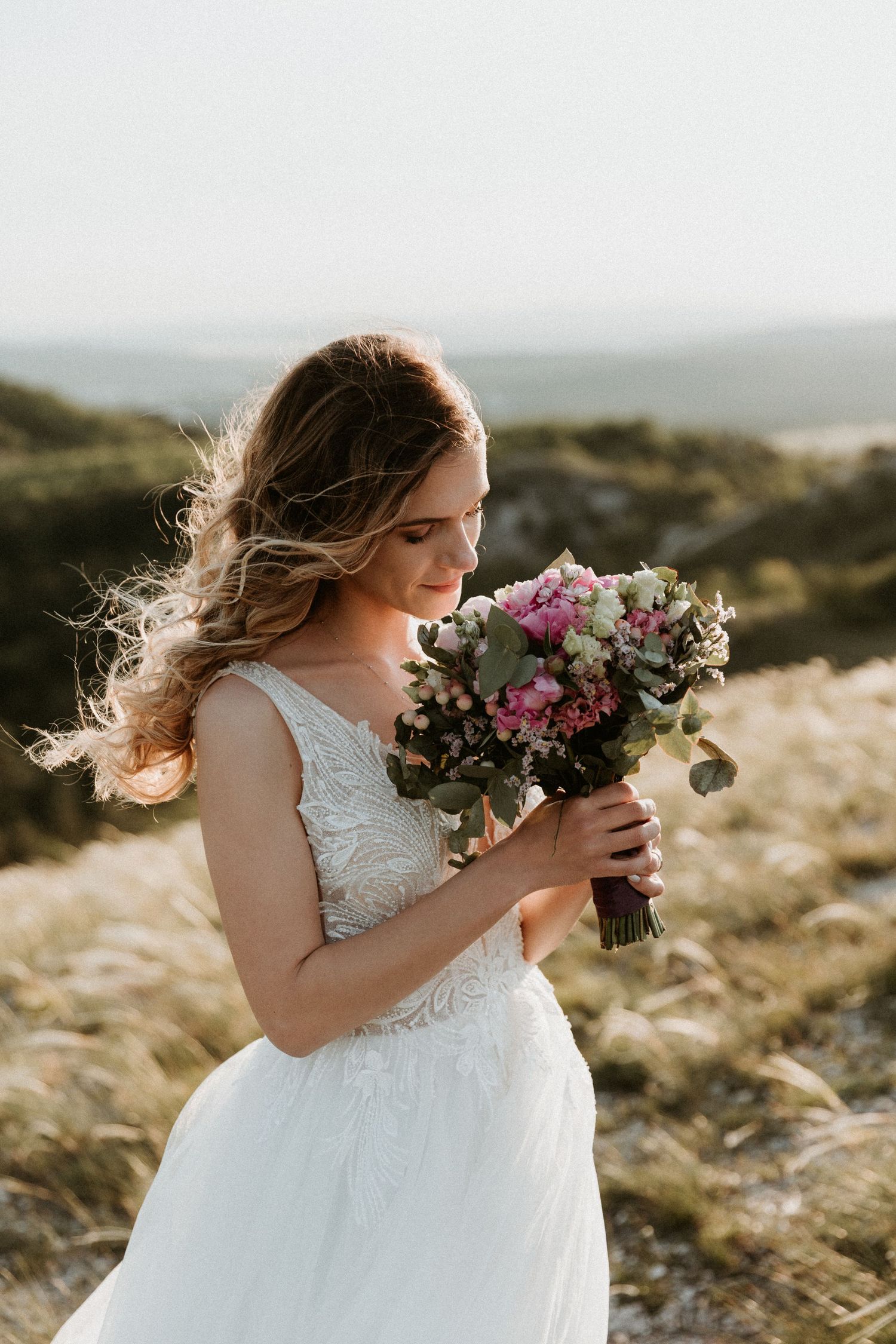 A bride in a white lace dress holds a colorful bouquet of pink and purple flowers against a sunset landscape.