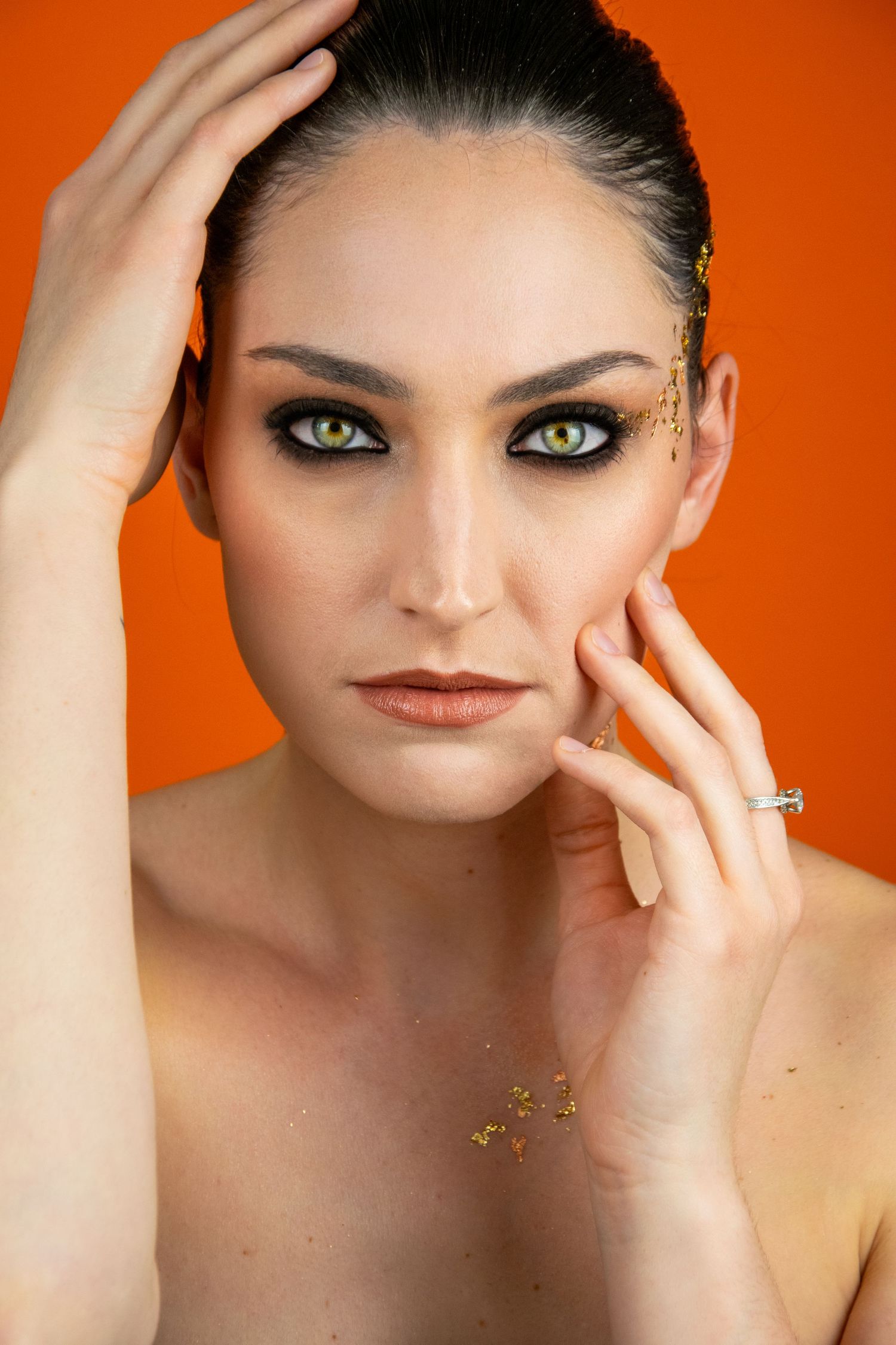 Beauty portrait against orange background with minimal makeup and gold accent details.