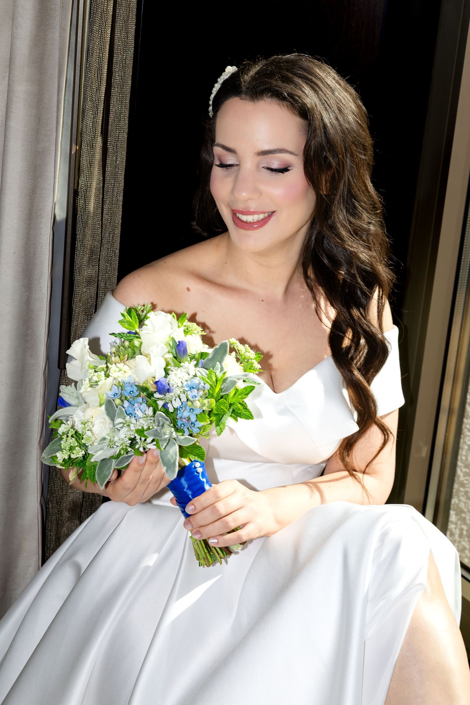 Smiling bride in white off-shoulder gown holding blue and white floral bouquet, seated by a window.