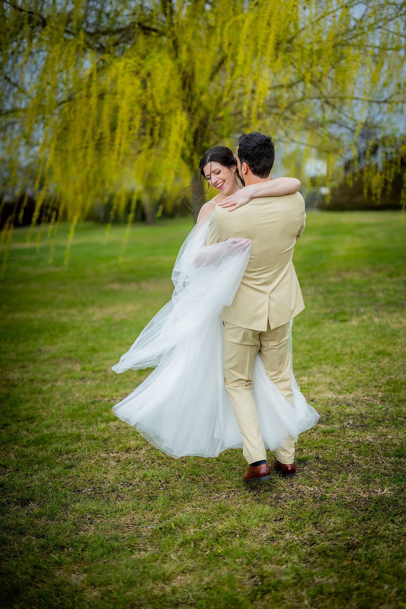 Groom in beige suit lifts smiling bride in white gown outdoors under a weeping willow tree on green grass.