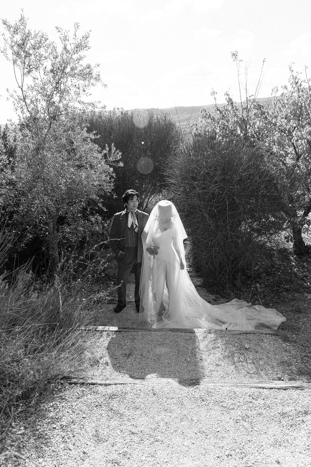 A black and white wedding photo showing a couple standing on a path surrounded by bushes and trees in Tuscany, Italy.