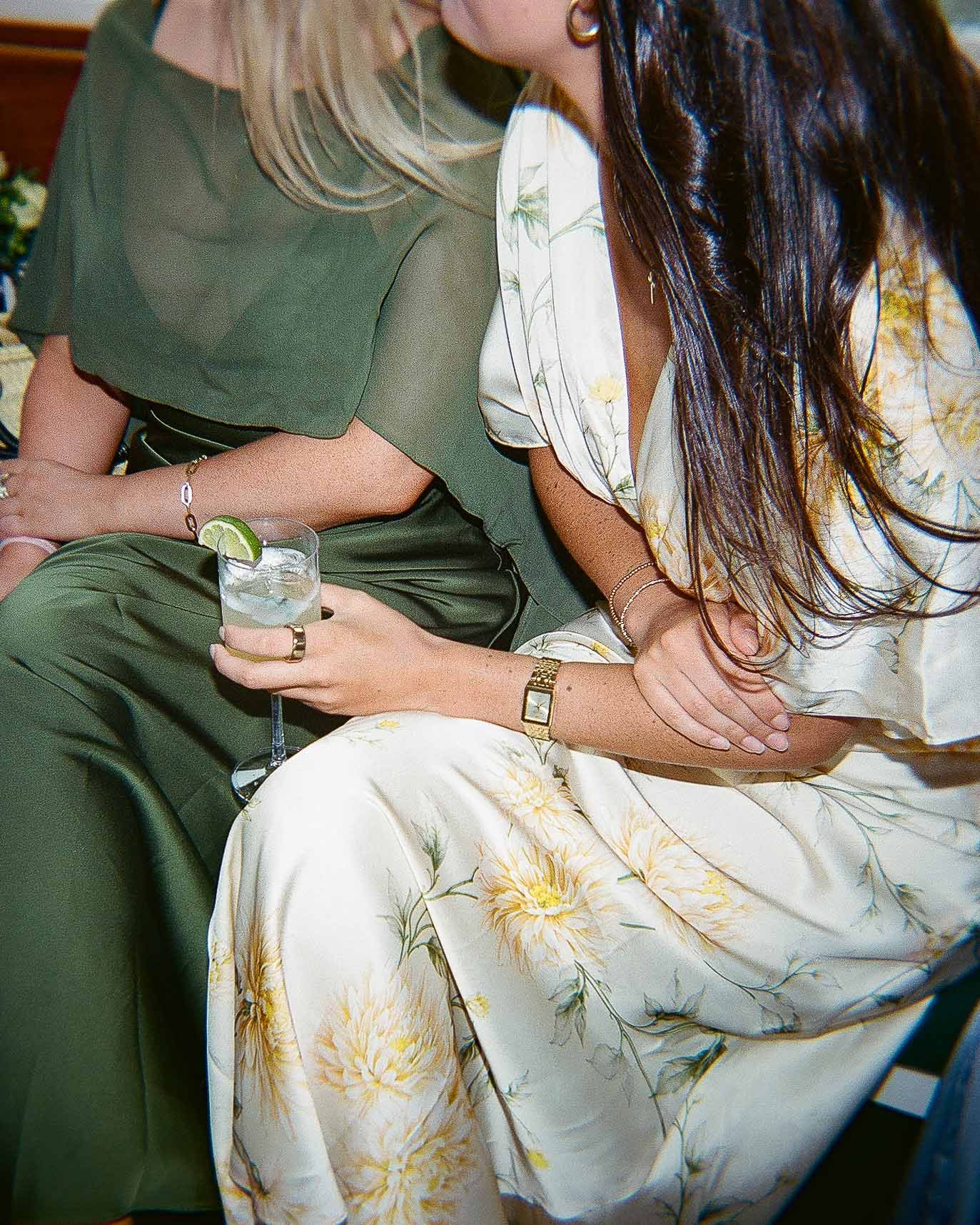 Two women sitting together, one in olive green dress holding cocktail with lime, other in white floral dress wearing gold watch.