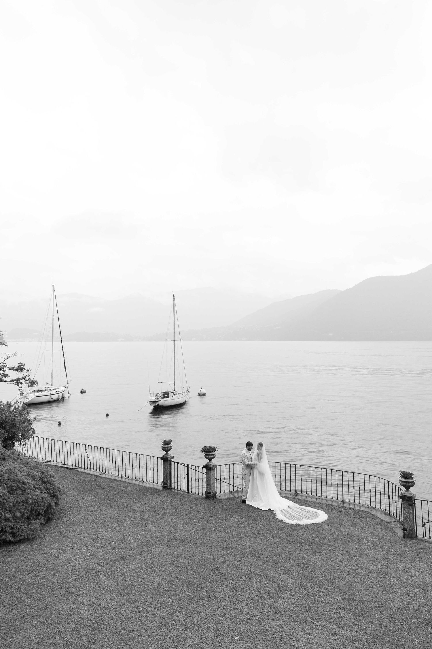 Black and white photo of bride and groom standing by lakeside fence with sailboats and mountains in background on Lake Como.
